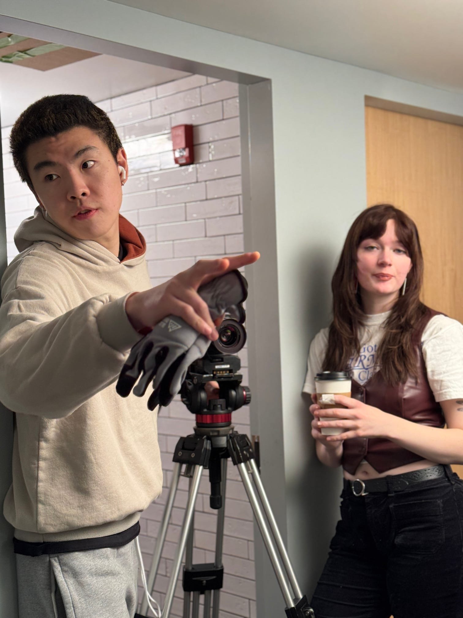 Two young women and a young man are in a hallway with gray brick walls, one man is taking a selfie with a camera on a tripod, the woman on the right is holding a coffee cup.