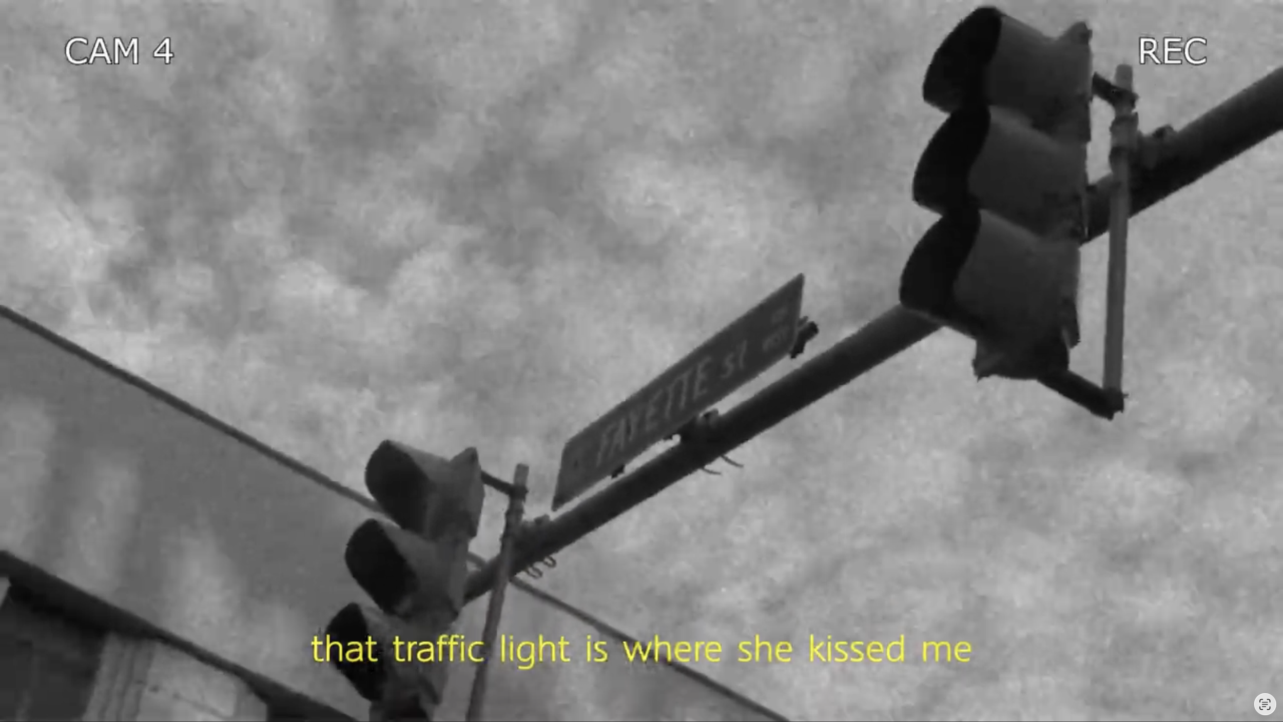 Black and white surveillance footage showing a traffic light at an intersection with a person standing nearby. The footage has timestamp and camera labels, and yellow subtitles saying "that traffic light is where she kissed me."