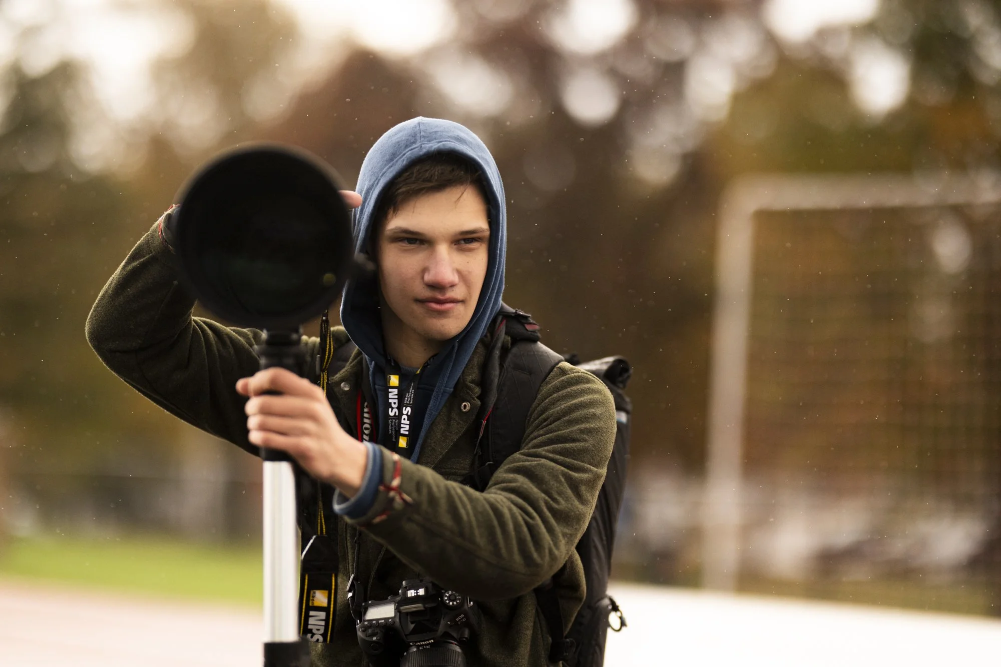 Young man in hoodie and jacket adjusting a tripod or camera equipment outdoors, with a blurred background of trees and a soccer goal.