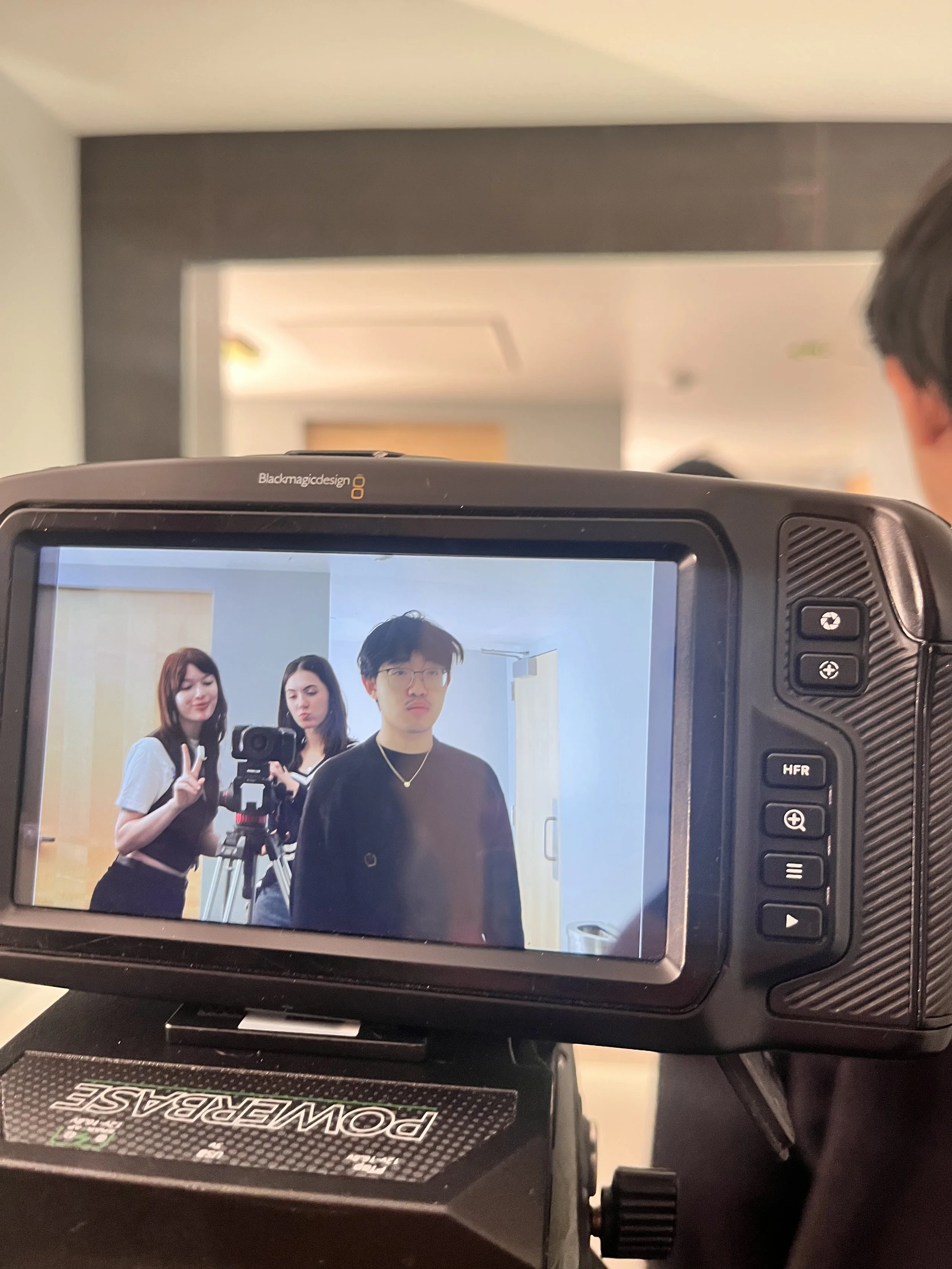 Three people taking a photo together with a camera in a room with light-colored walls and a doorway. The person in the foreground is wearing glasses and a black shirt, while the two women behind him are making peace signs and looking at the camera. T