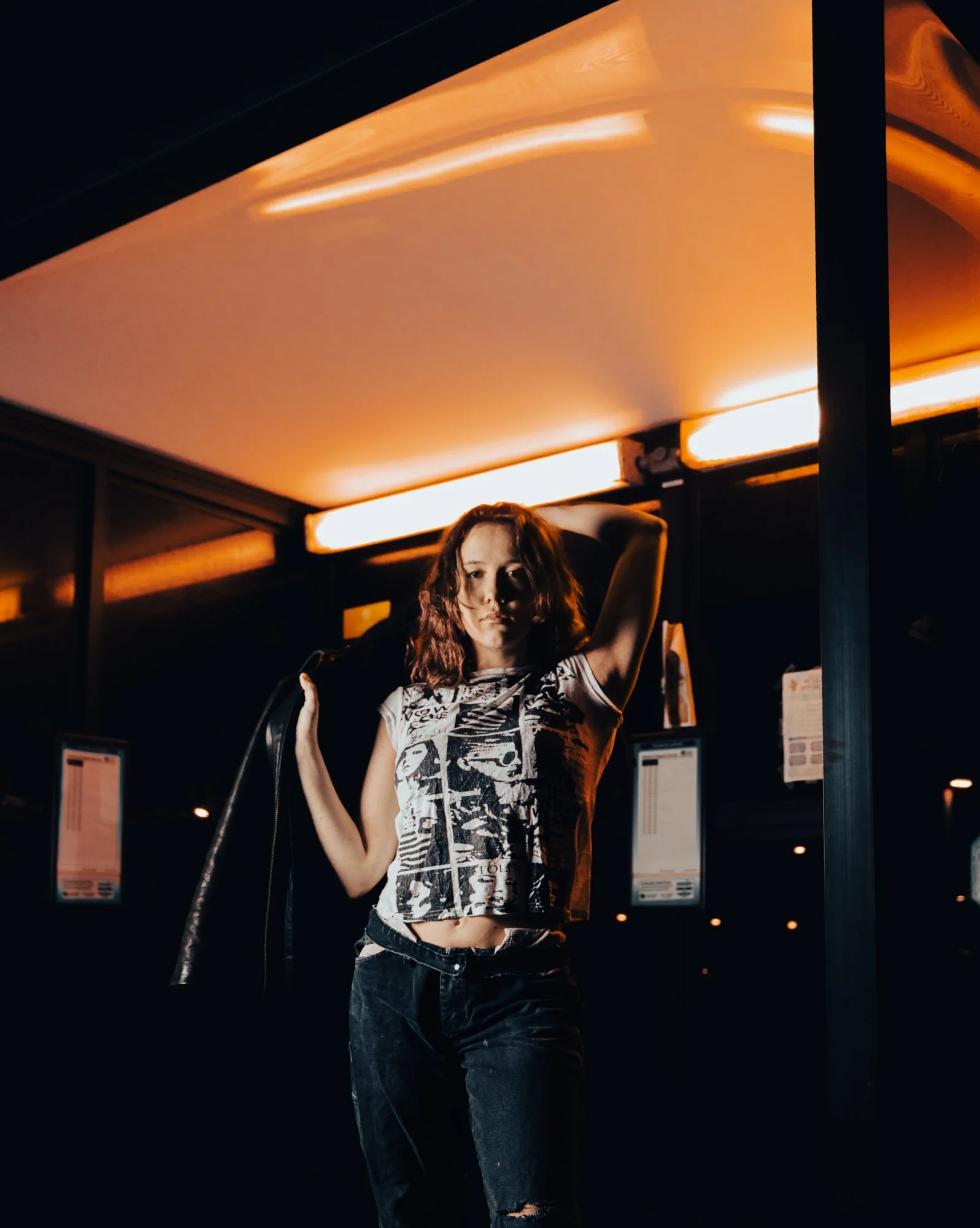 A young woman with long wavy hair standing in a dimly lit environment with orange lighting, crossing her arms and wearing a graphic t-shirt and ripped jeans.