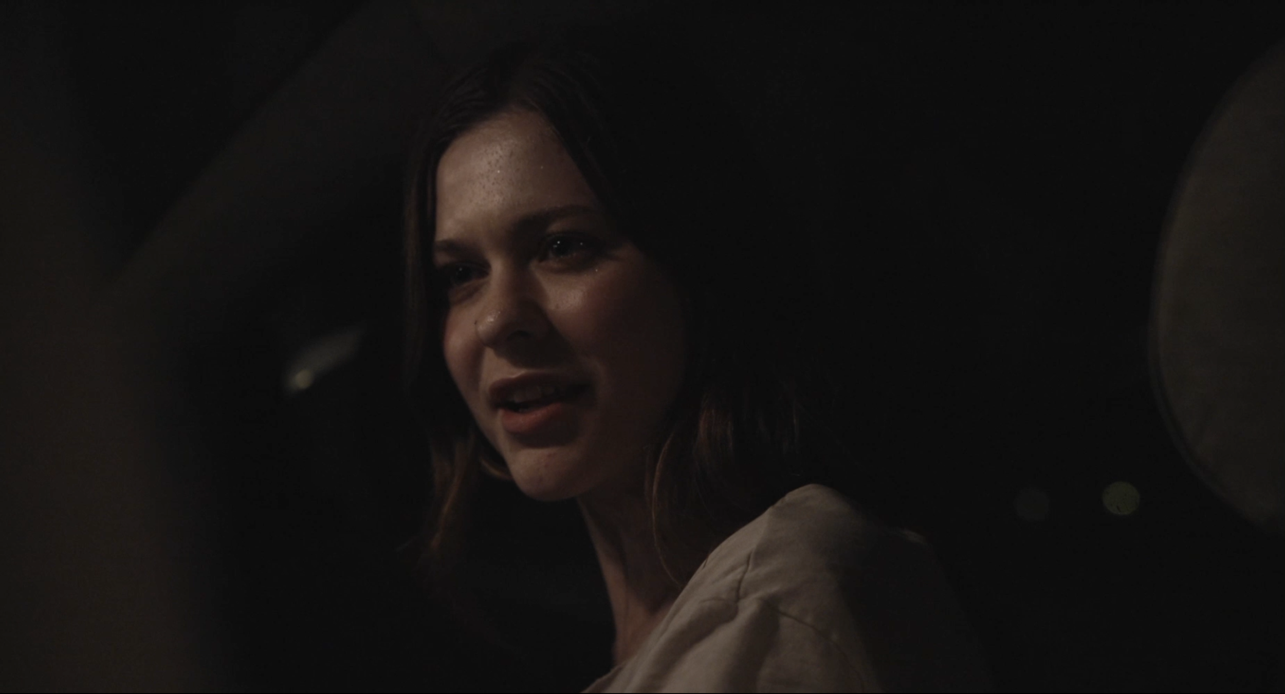 A woman with brown hair is seen through a circular opening, looking to her left with an expression that may suggest surprise or concern, in a dimly lit environment.