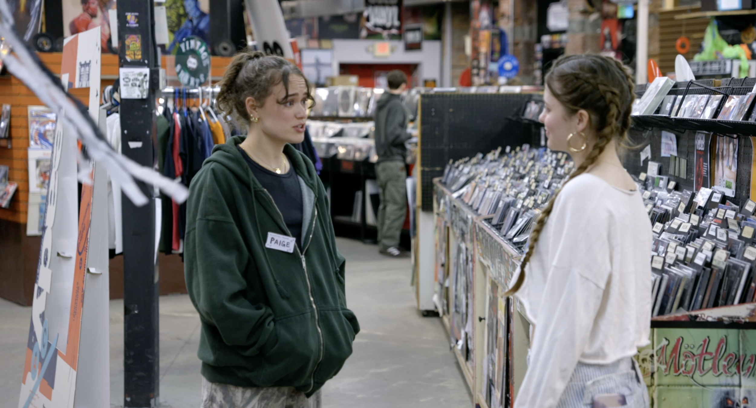 Two women standing and talking in a record store, with shelves of CDs and DVDs behind them.