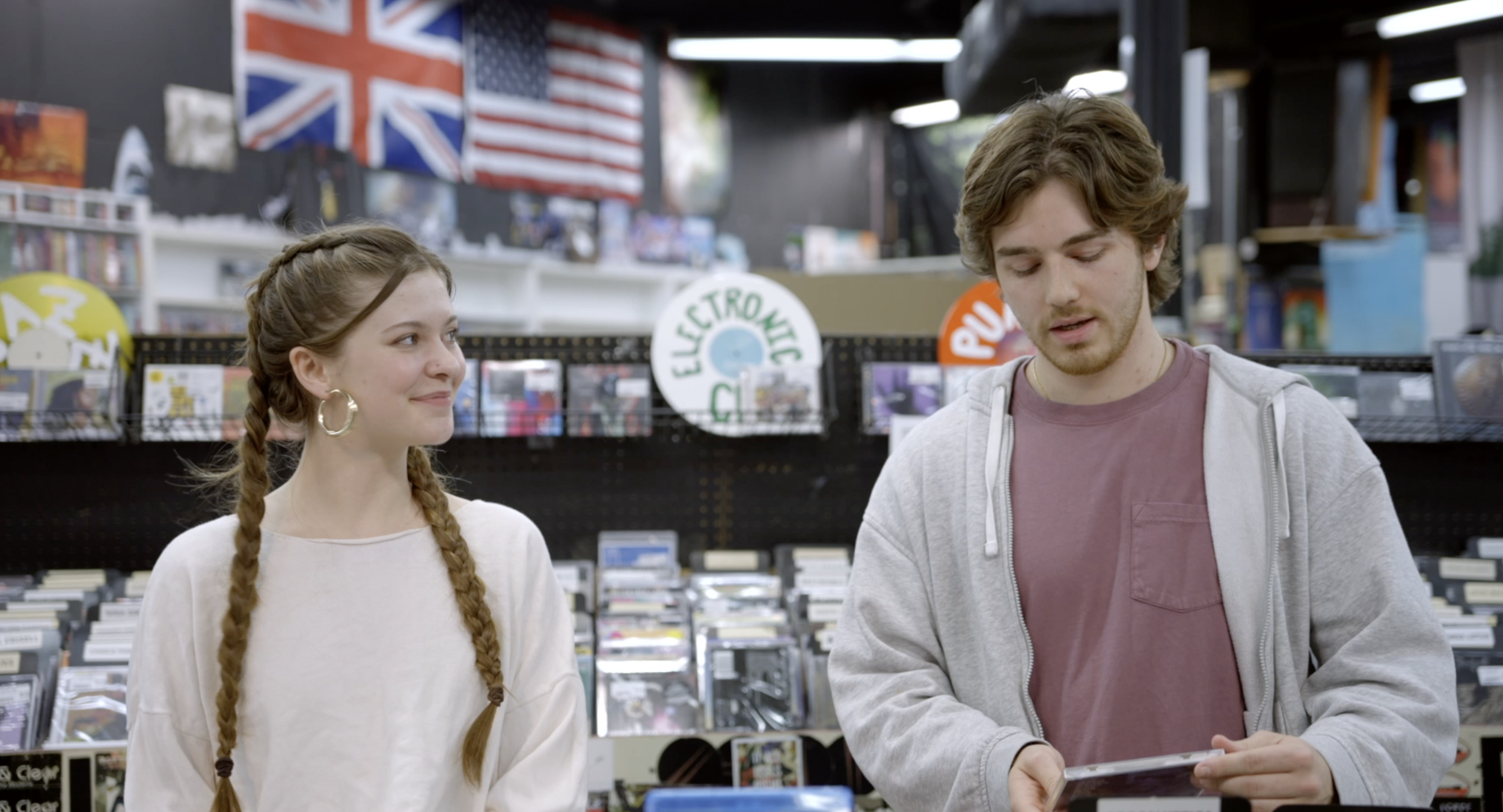 A young woman with long braided hair and hoop earrings smiling at a young man with wavy hair, wearing a gray hoodie, as they browse items in a record store with British and American flags hanging on the wall behind them.