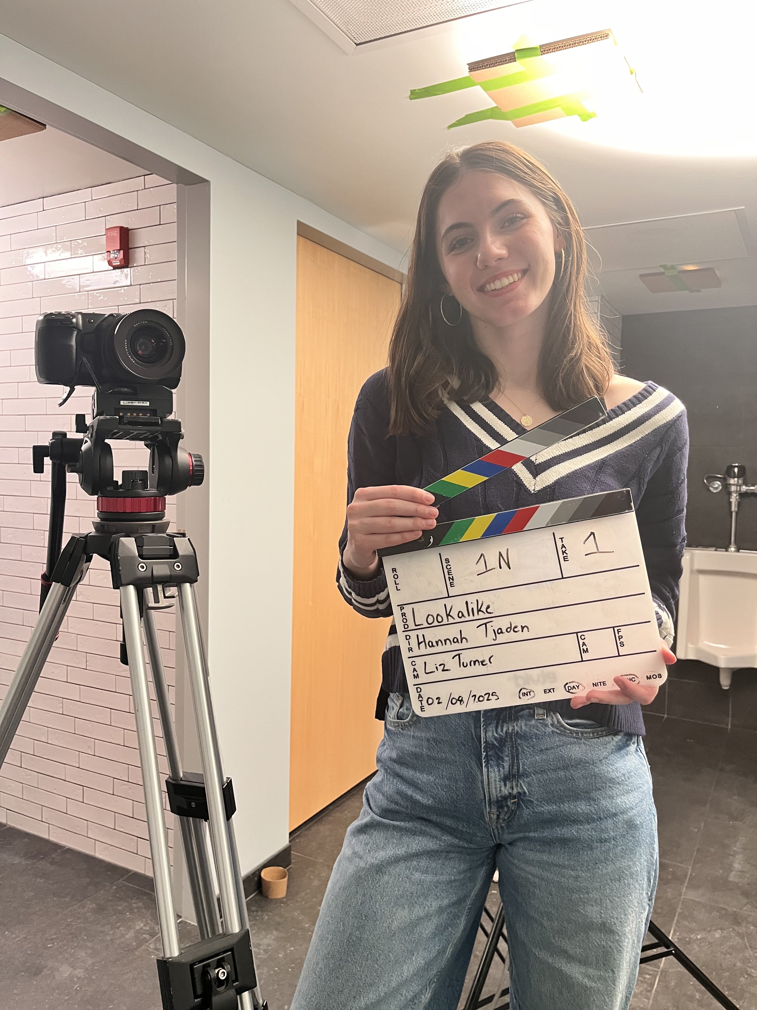 A young woman smiling and holding a film slate with names written on it, standing in front of a camera on a tripod for a film or video production.