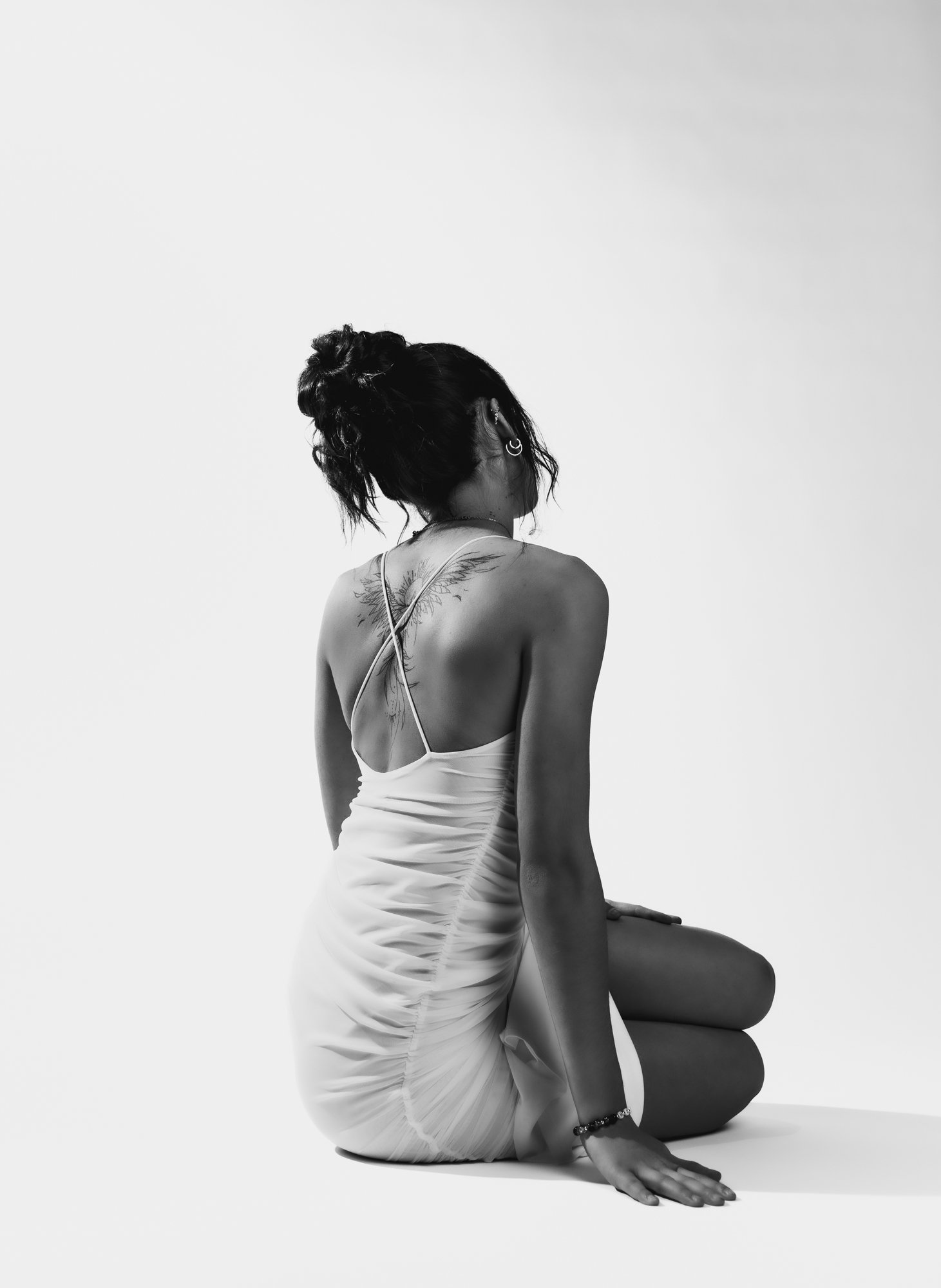 A woman dressed in a light-colored dress is sitting on the floor with her knees bent and her head turned to the side, facing away from the camera. She has dark hair styled in a messy bun and tattoos on her back and arm. The background is plain and wh