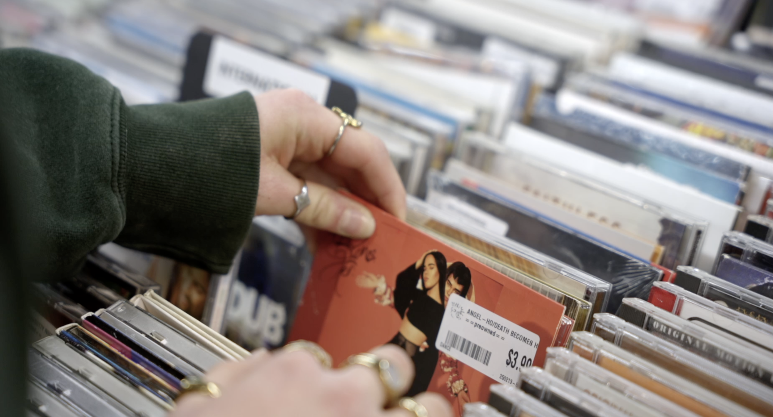 A person with rings on their fingers browsing through CDs in a store, with shelves of music in the background.