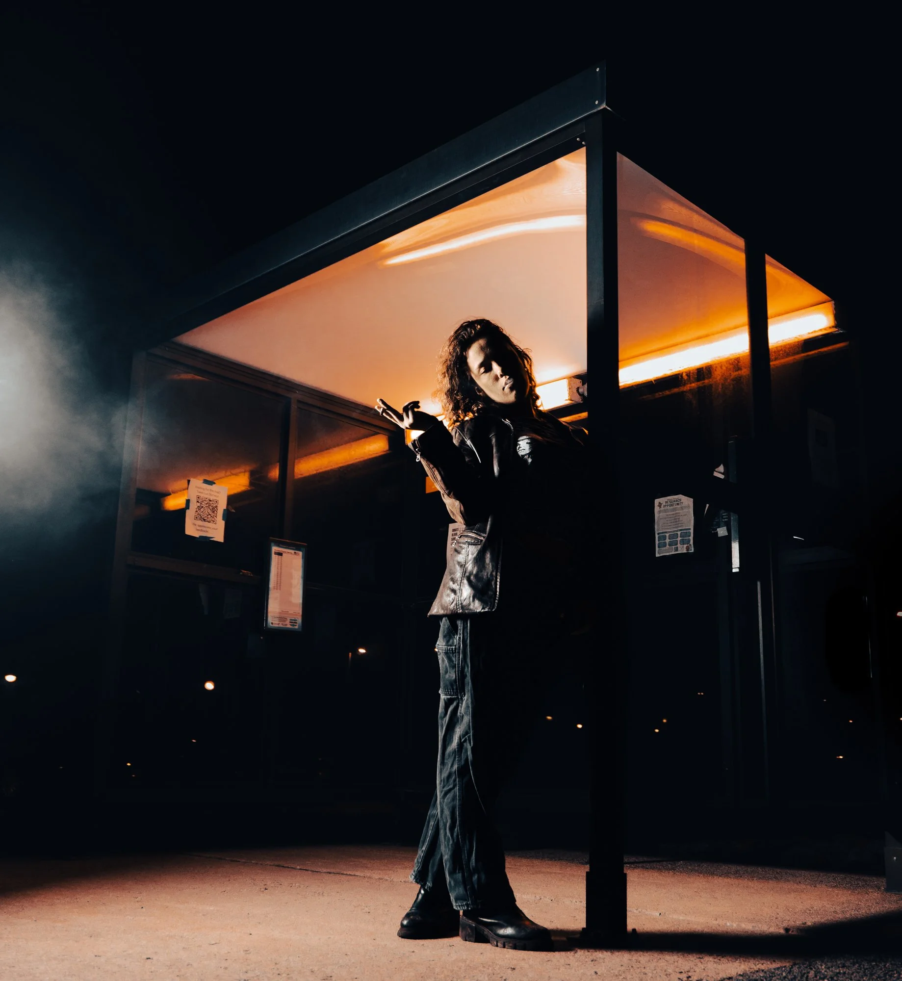A person with curly hair and dark clothing standing in a dark, minimalistic environment under an orange-lit structure, with one hand raised and the other by their side, in front of a dark sky.
