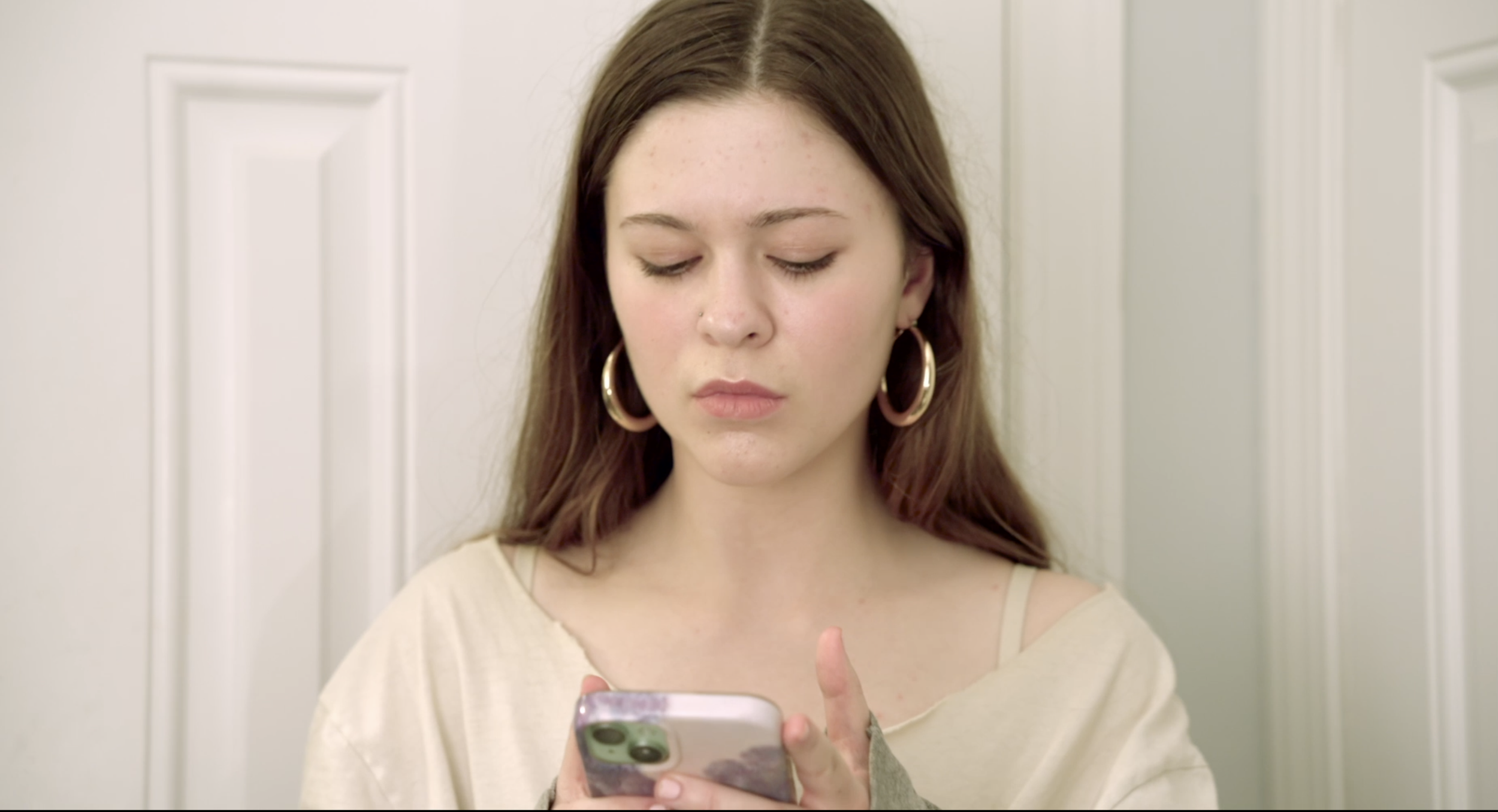 A young woman with long brown hair and large hoop earrings looking at her smartphone, standing in front of a white paneled door.