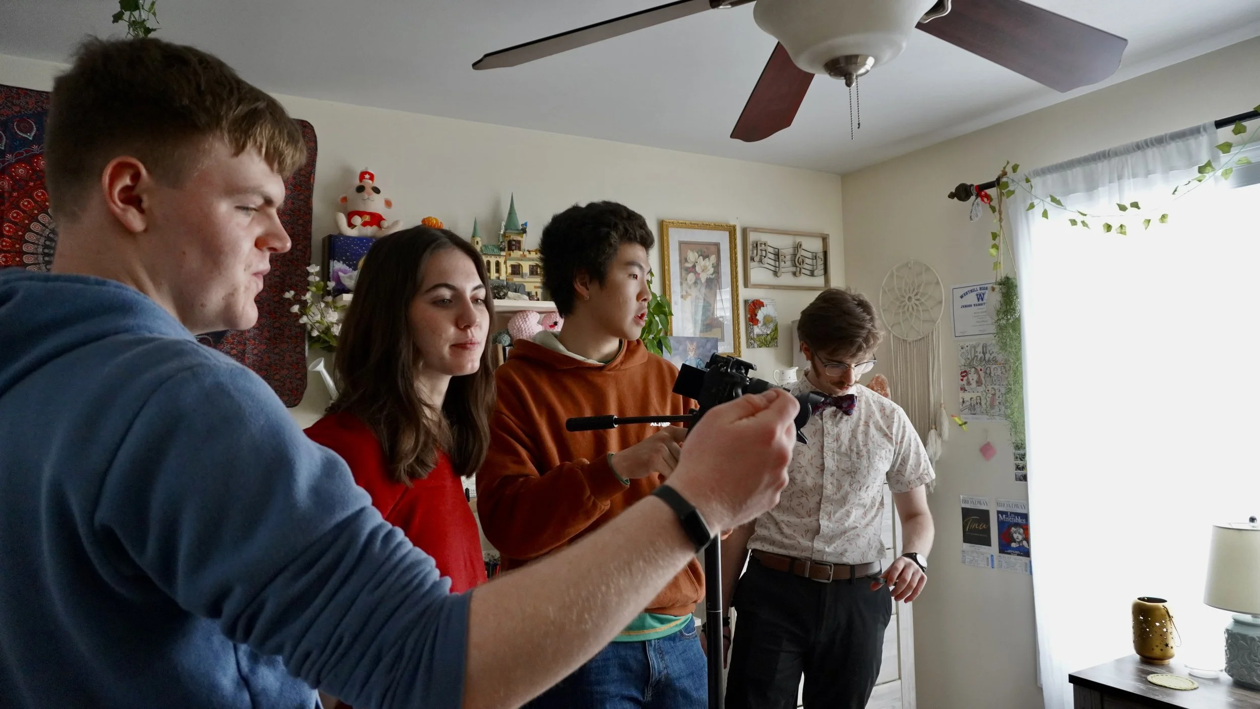 Group of five young people standing indoors, one holding a camera on a tripod, other four looking at the camera or the person holding it, in a room decorated with various pictures, plants, and ornaments, including a stuffed animal on a shelf.