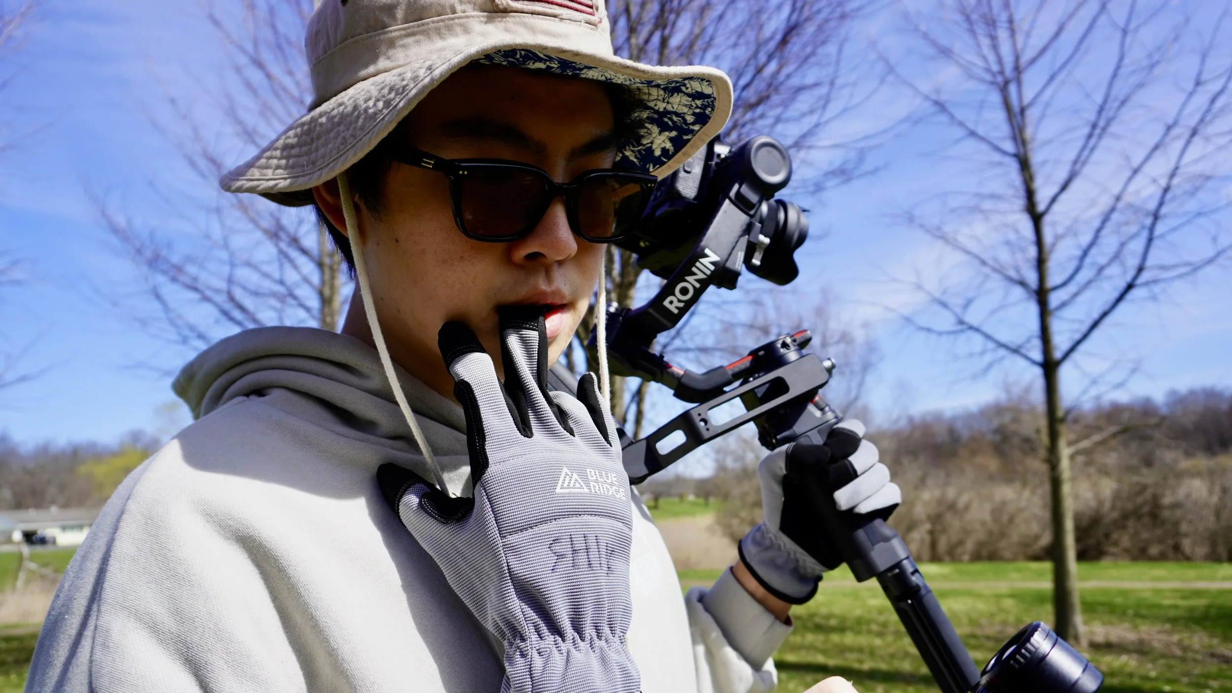 A person wearing sunglasses, a wide-brimmed hat, gloves, and outdoor clothing holding a camera tripod outdoors on a sunny day with trees and a park in the background.
