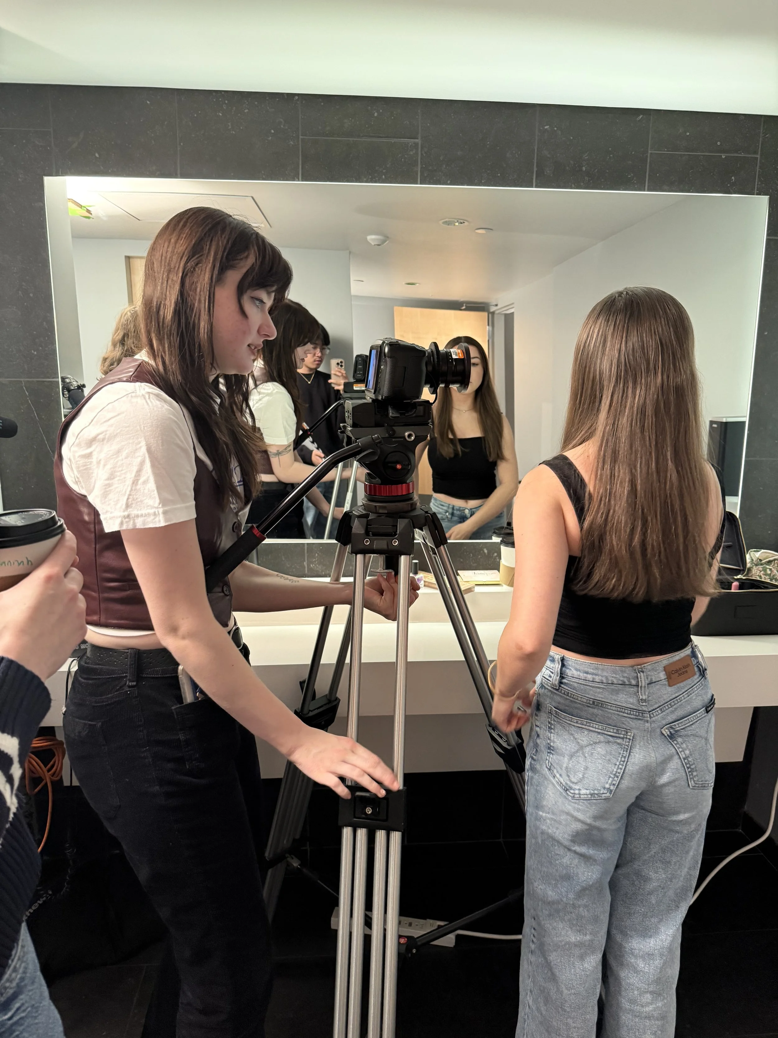 Group of women in a bathroom taking photos and preparing for a photo shoot, with a camera on a tripod facing them.