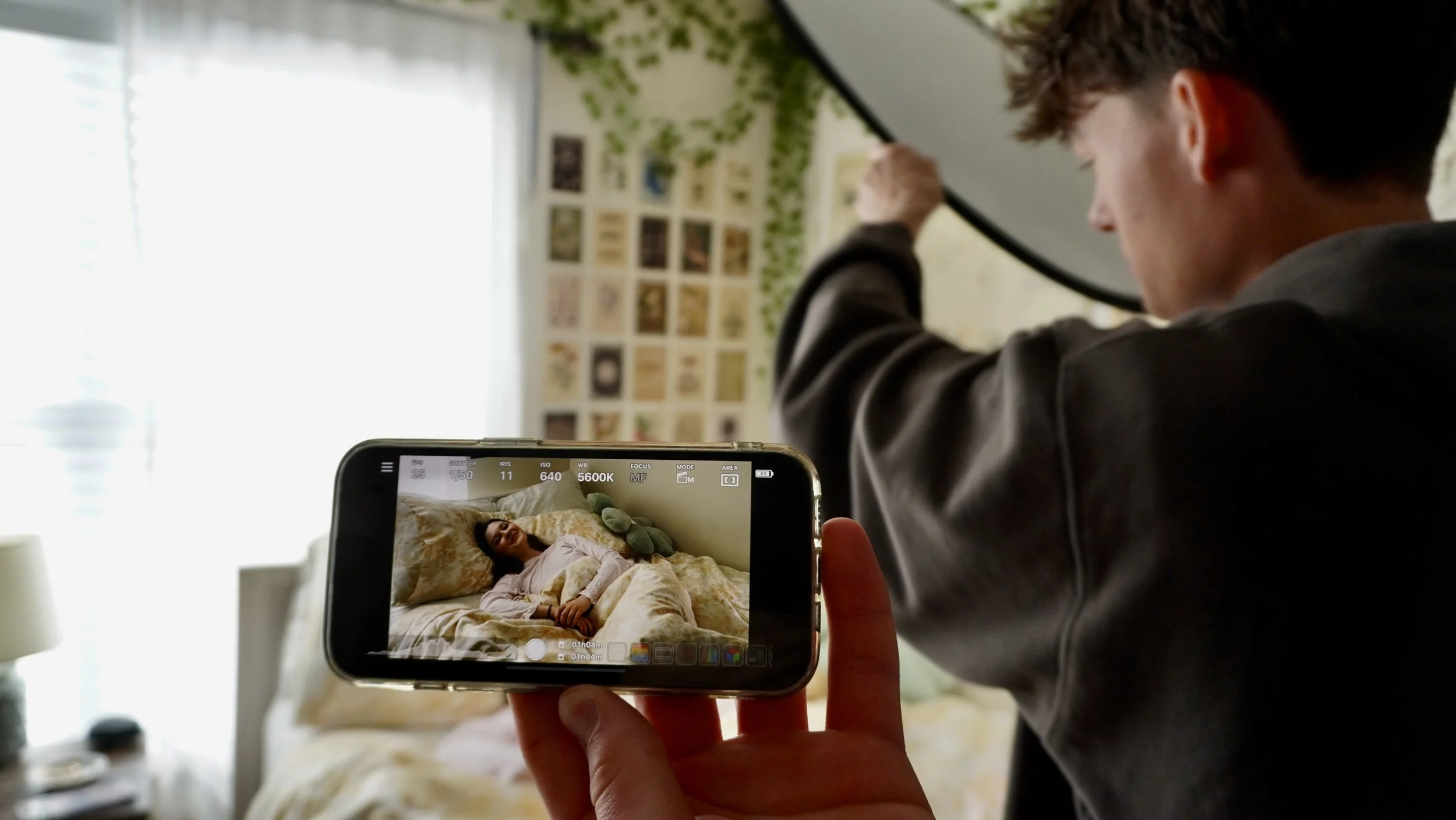 Person in gray hoodie taking a photo of a woman lying in bed with a smartphone, with the phone's screen displaying the woman smiling and resting against pillows.
