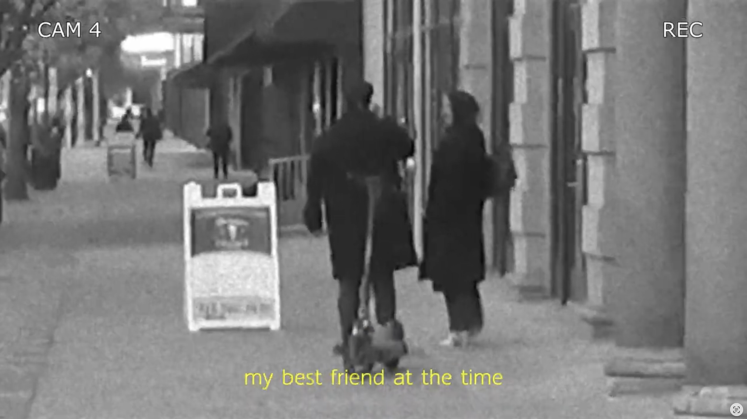 Two women walking together on a city sidewalk, with other pedestrians in the background. The women are engaged in conversation, and there is a signboard on the sidewalk. The scene is in black and white with subtitles at the bottom reading 'my best friend at the time.'