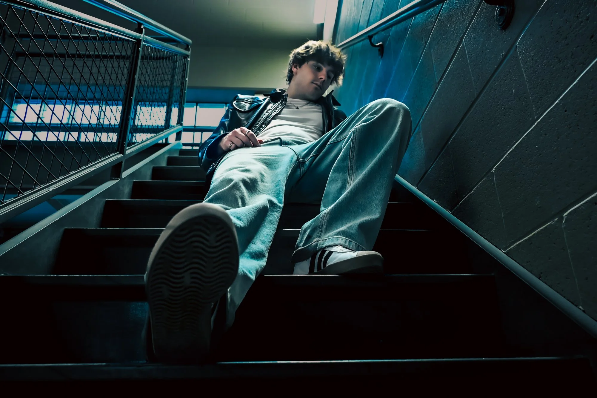 Young person with curly hair, wearing a leather jacket and light-colored pants, sitting on stairs in a building with geometric wall patterns, looking down with a relaxed pose.