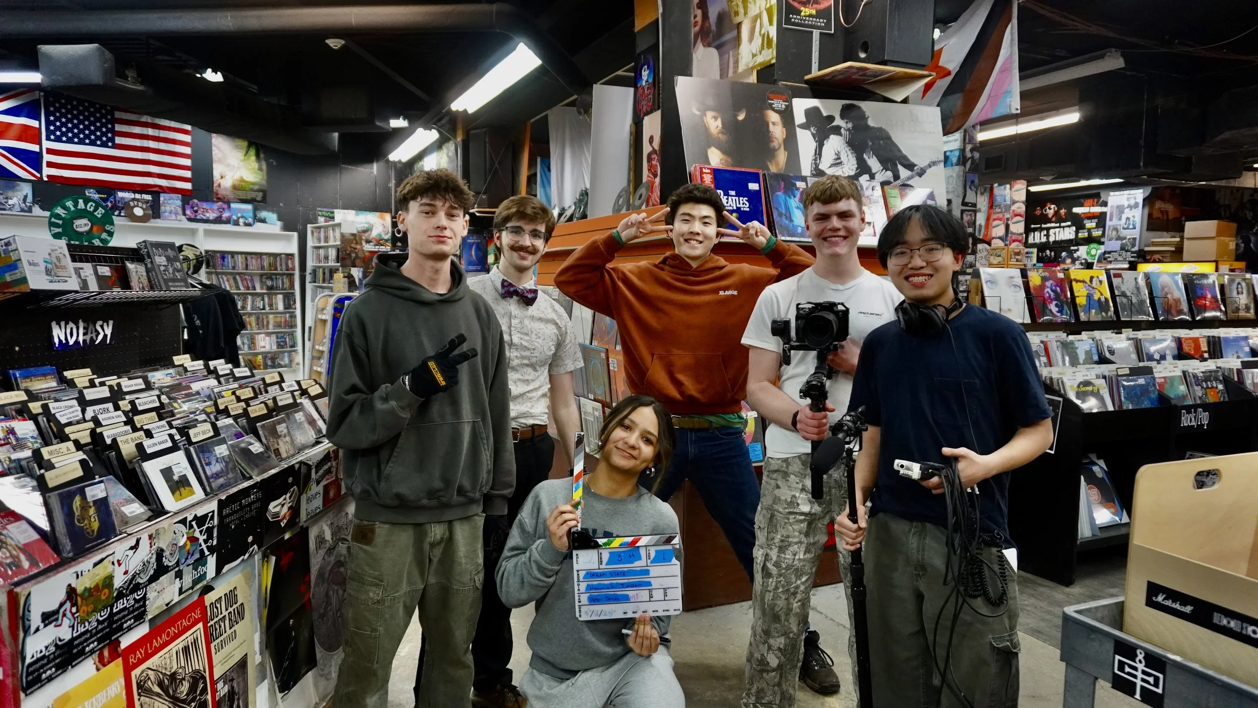 Group of seven diverse young adults standing in record store with music posters, flags, and shelves of CDs and vinyl. They are smiling and holding various filming equipment.