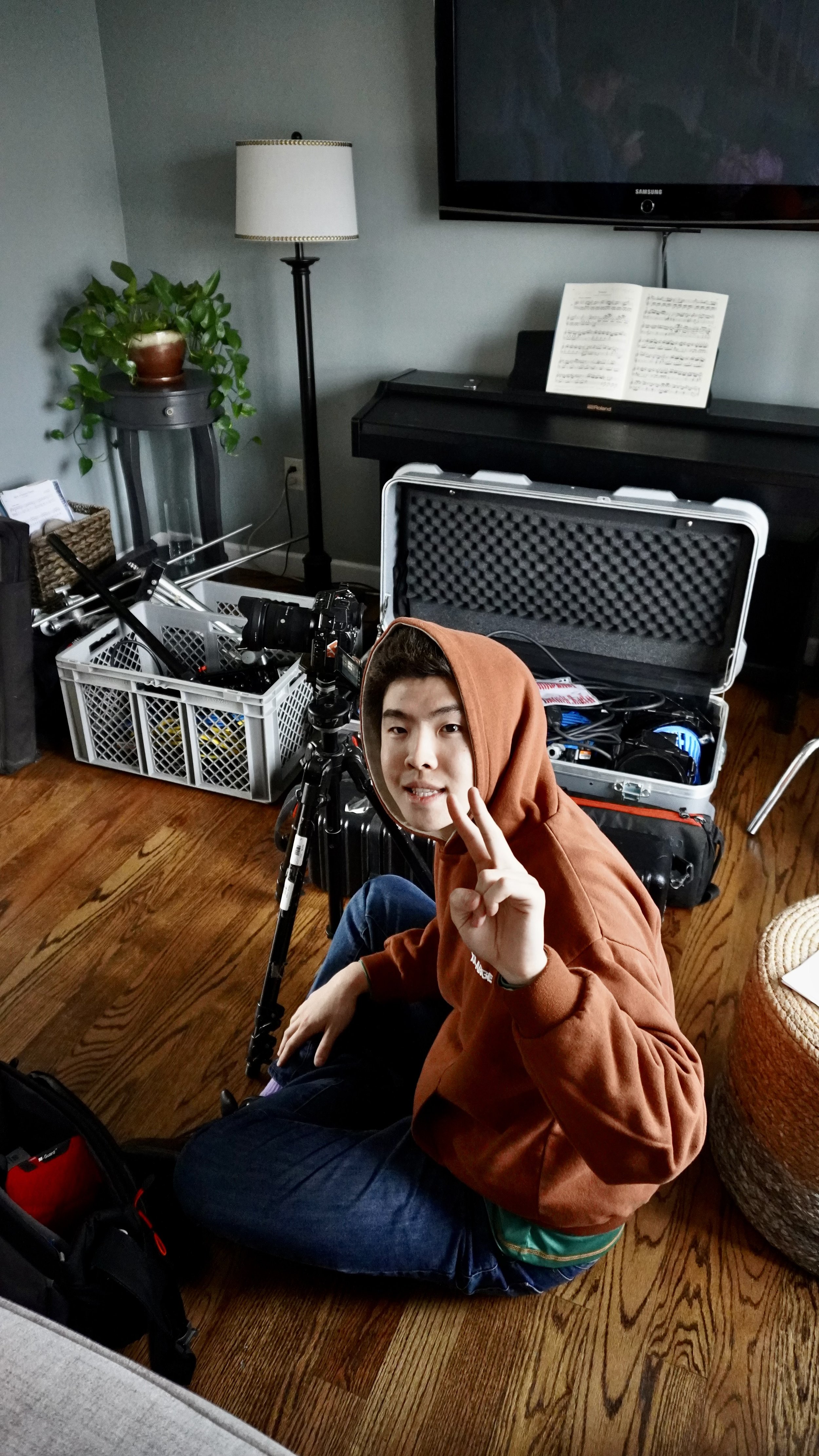 A young man with a hoodie sitting on a hardwood floor, smiling, with a peace sign, surrounded by camera equipment and a music stand with sheet music in a room.