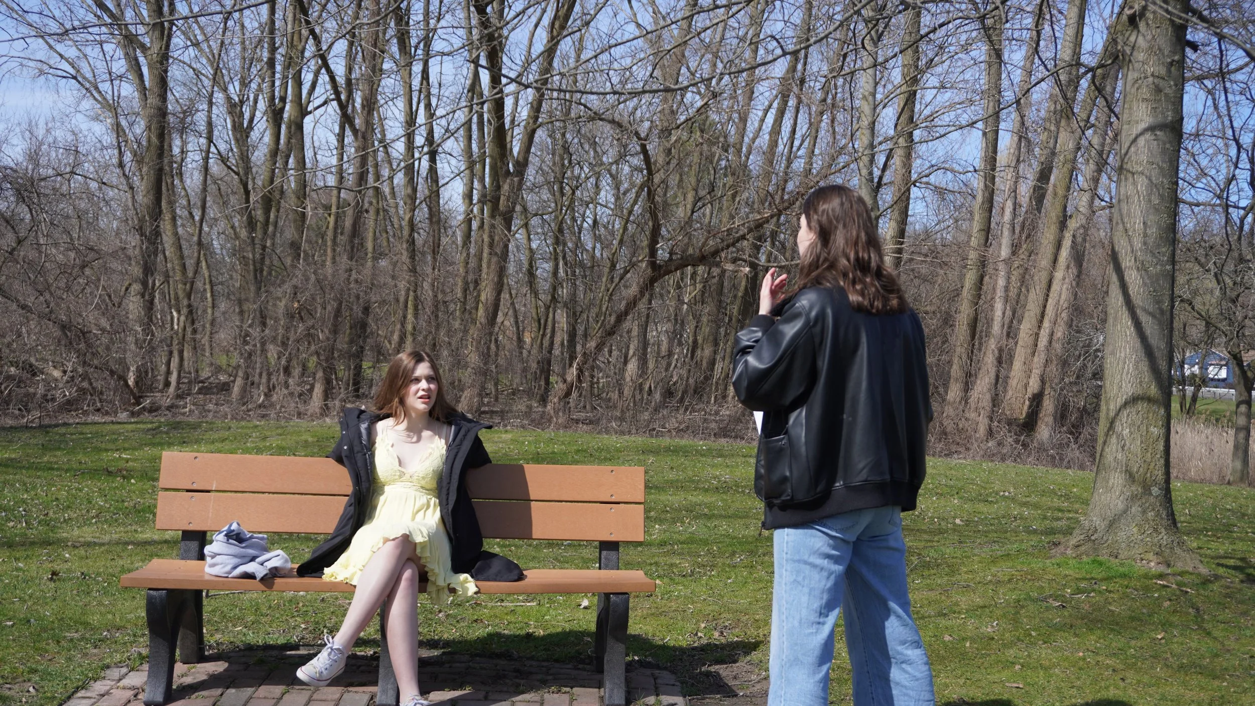 Two women converse outdoors near a wooden bench in a park with leafless trees and a blue sky.