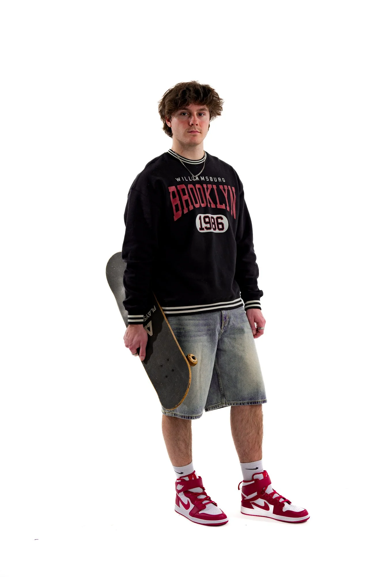 Young man standing with one hand in his pocket, holding skateboard with the other, wearing a black Brooklyn sweatshirt, denim shorts, white socks, and red and white Nike sneakers, with a chain necklace, against a white background.