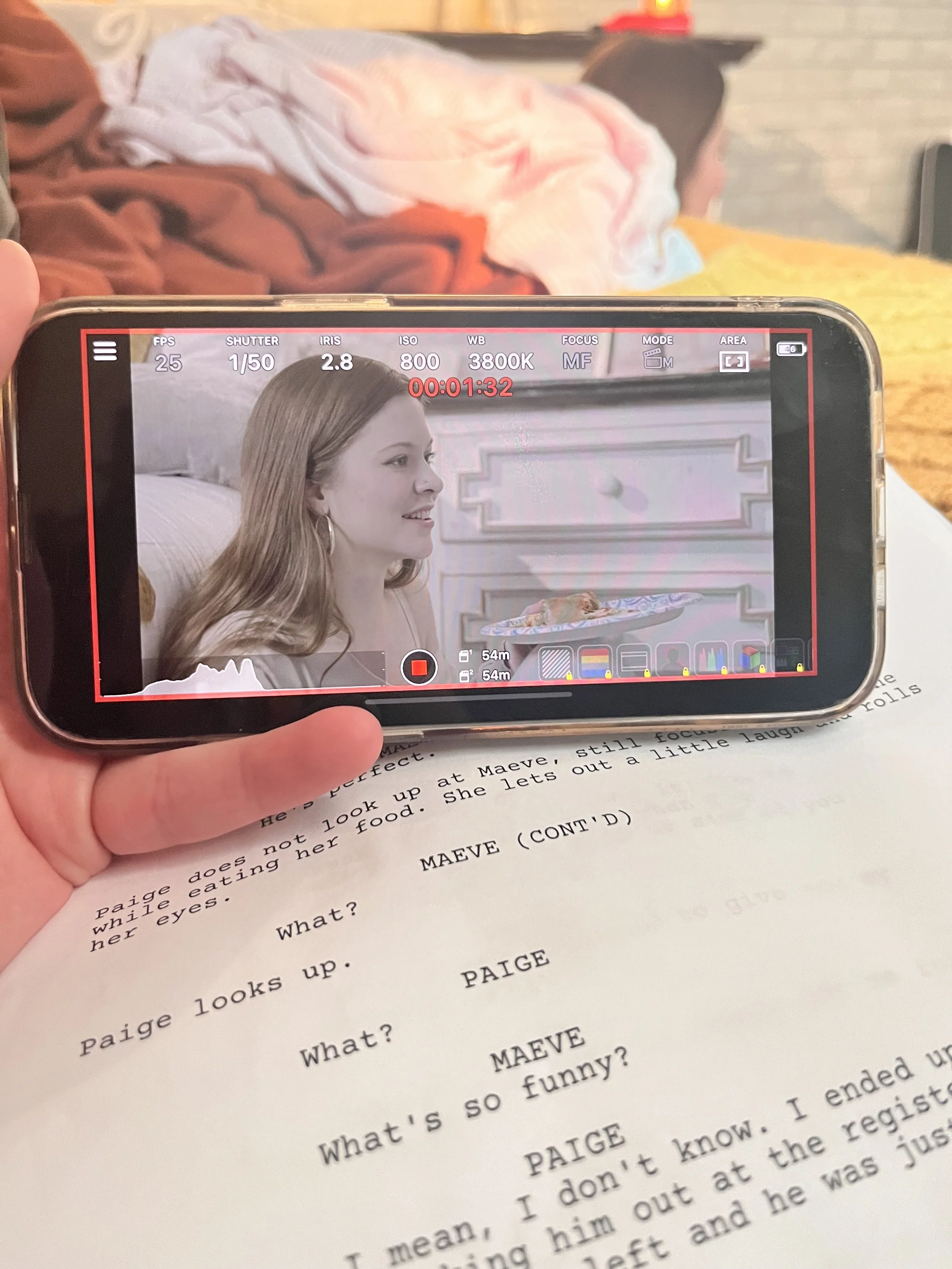 A person holding a smartphone capturing a photo of a woman smiling, sitting at a table with printed text in front of her. The background shows a books or shelves and a yellow cloth.