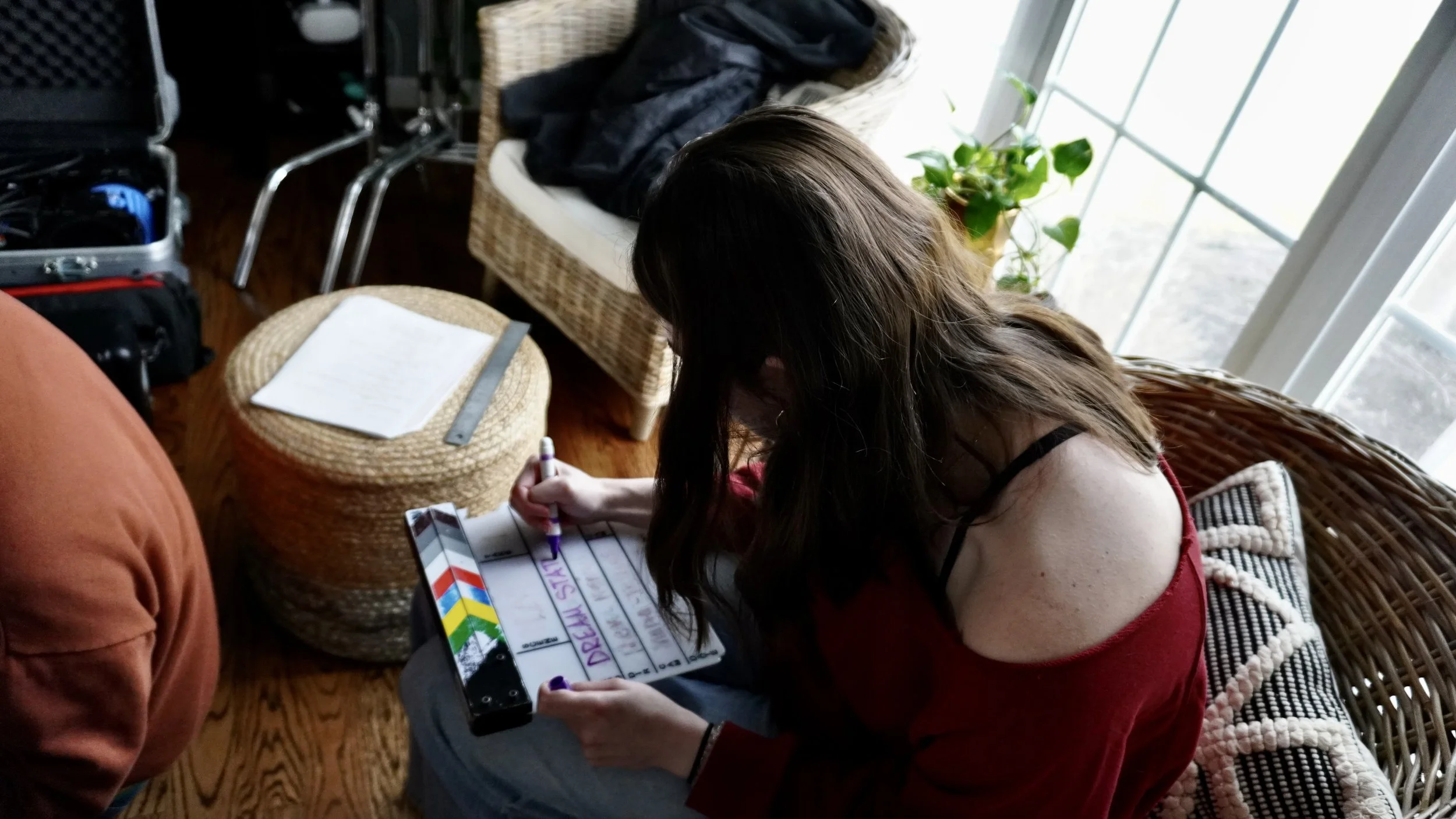 A woman with long brown hair sitting on a wicker sofa near a large window, writing on a clipboard with a marker. There are potted plants and a wicker ottoman with a ruler and paper on it. Items are scattered around, including a blanket and a black ja