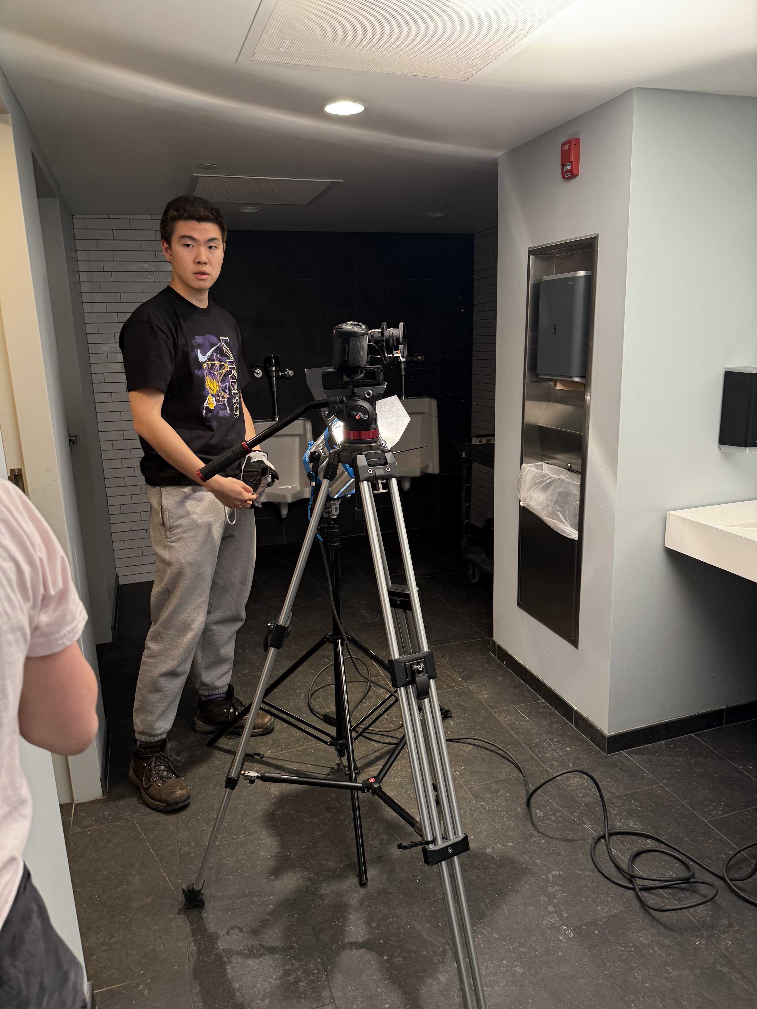 A young man standing next to a camera on a tripod in a bathroom, with a sink, mirror, and wall-mounted items visible.