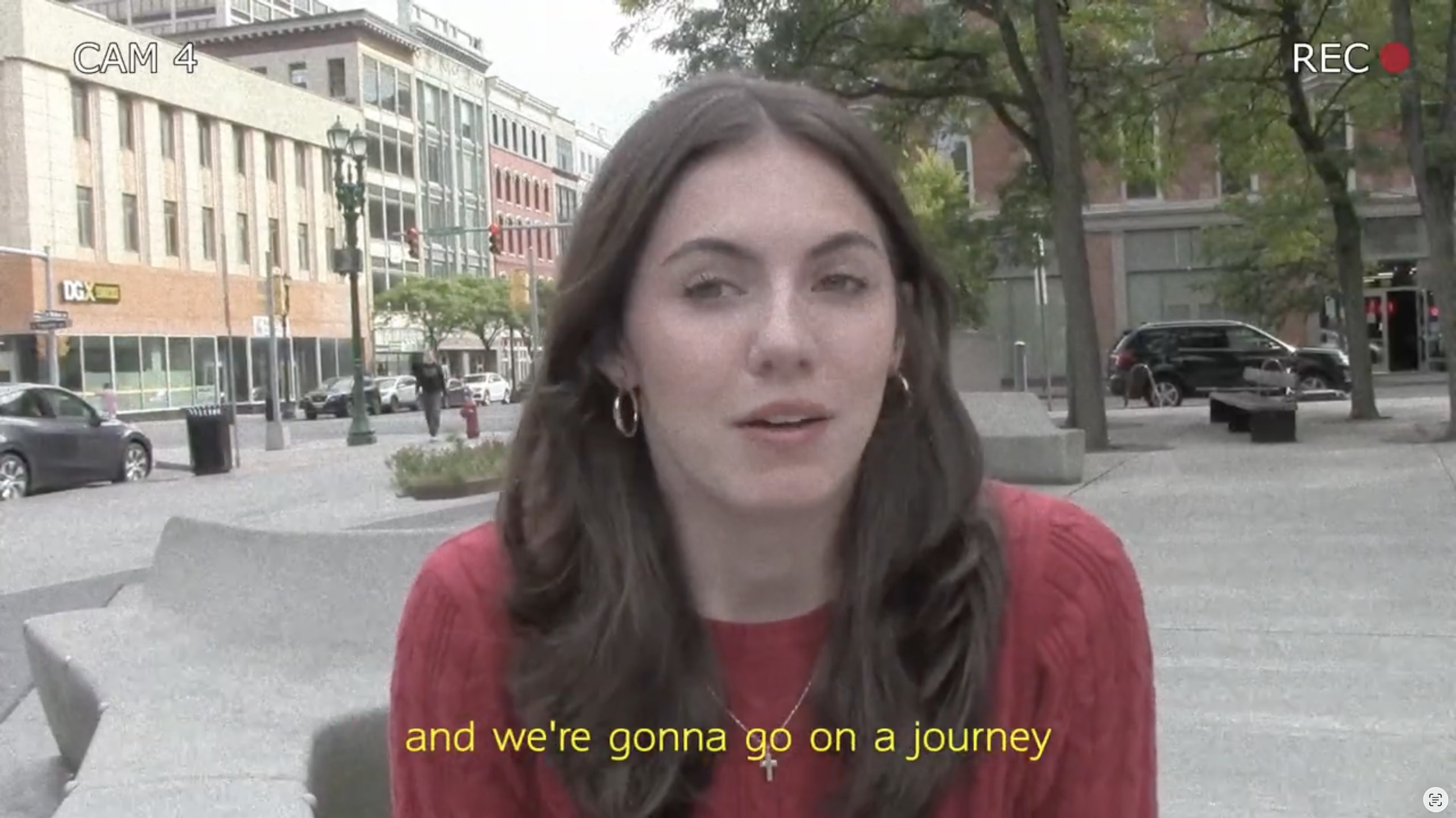A woman with long brown hair and hoop earrings wearing a red top is sitting on a concrete bench outdoors in an urban park or plaza, with buildings, trees, and parked cars in the background. She appears to be speaking, with subtitles reading 'and we're gonna go on a journey.'