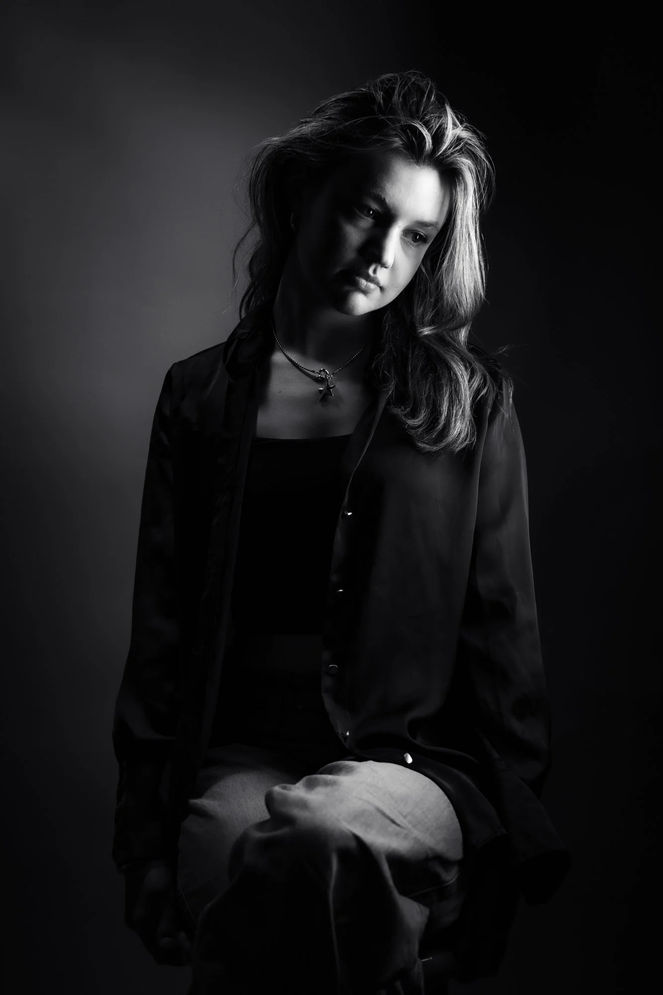 Black and white portrait of a young woman with long wavy hair, wearing a dark jacket and a necklace with a star pendant, seated with her hands on her lap, looking downward with an introspective expression, against a dark background.