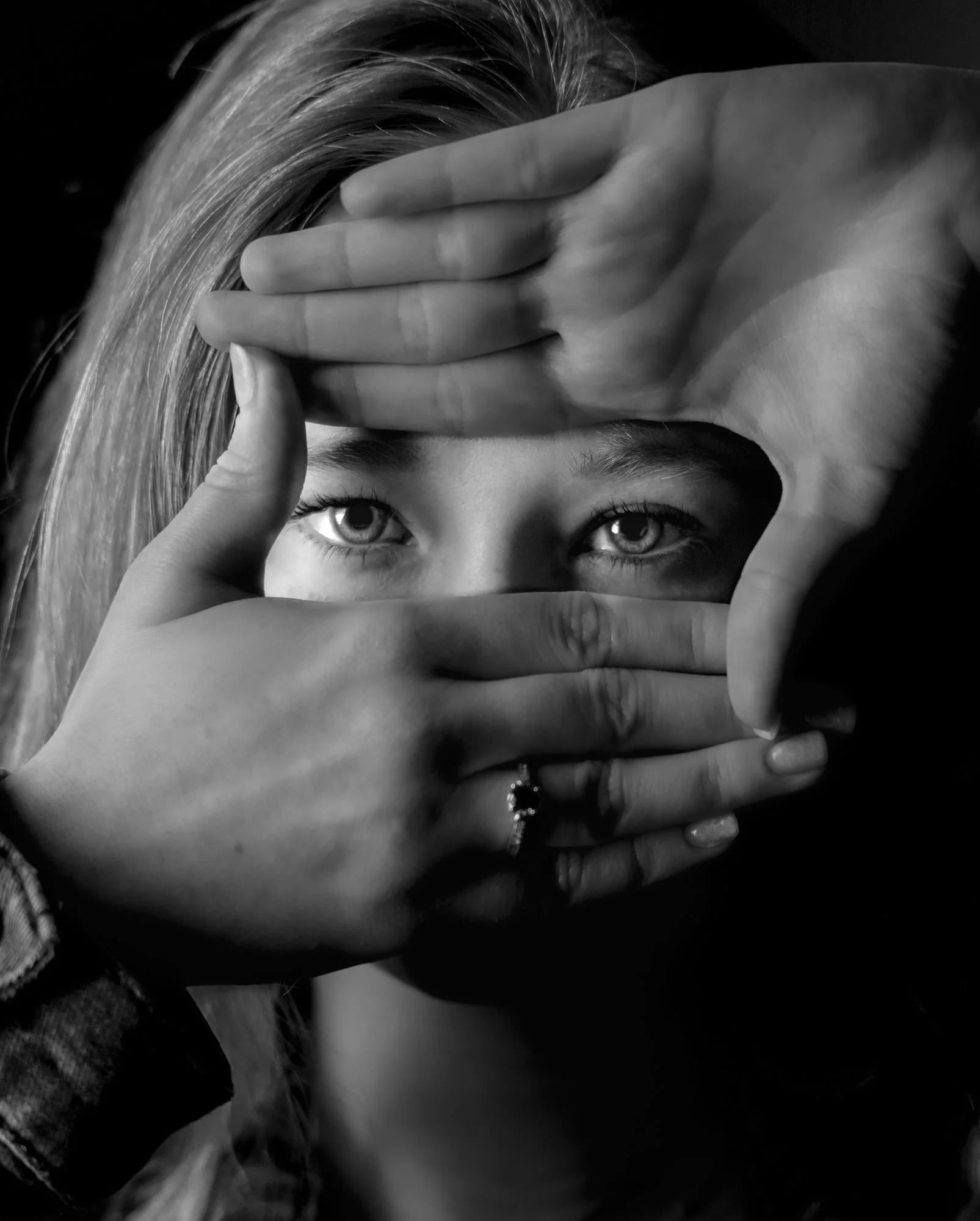 A person with light-colored hair holding their hands in front of their face, creating a frame that highlights their eyes, which are looking directly at the camera.