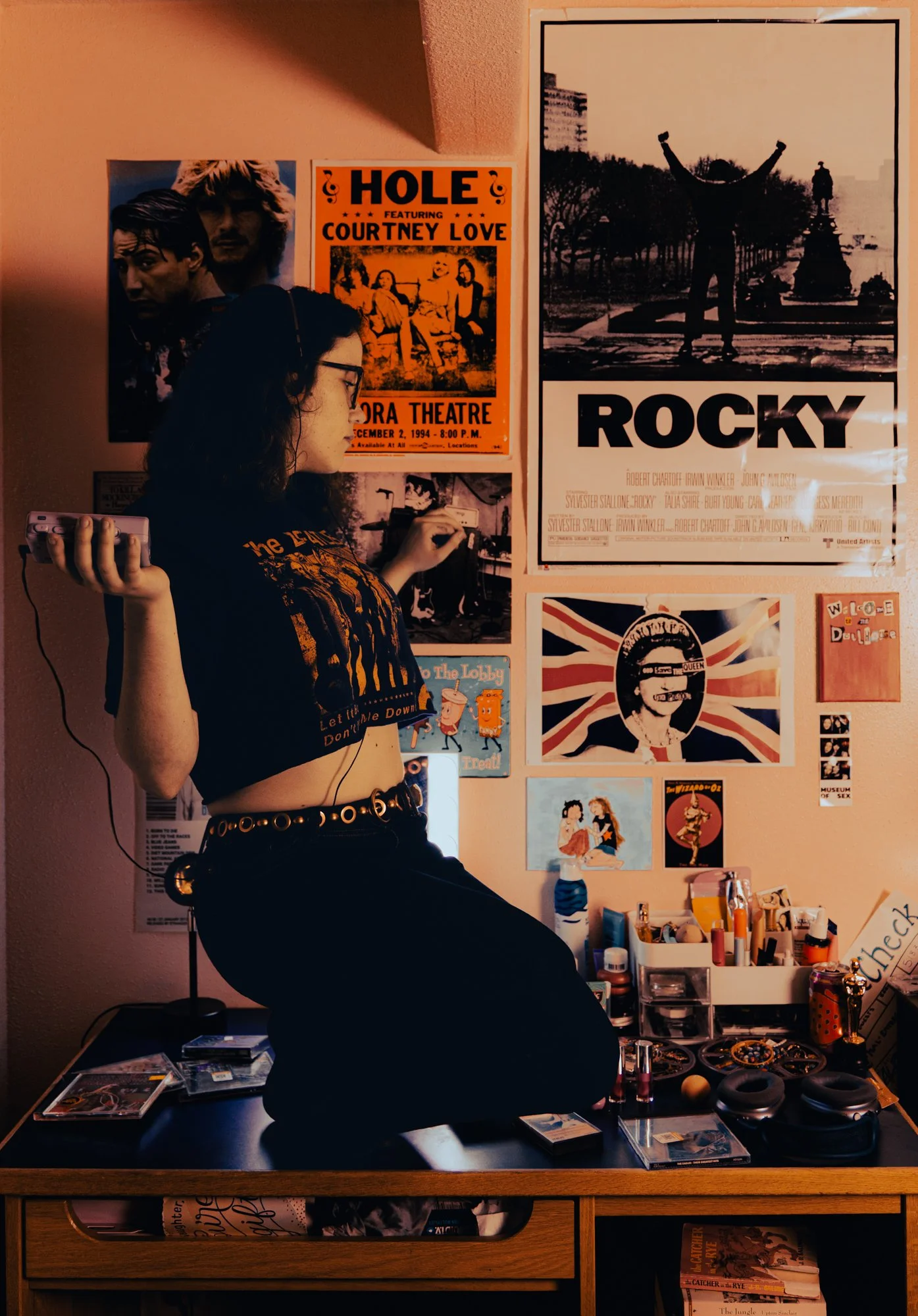 A young woman with dark hair and glasses is sitting at a desk with makeup items, CD cases, and a gaming console controller. She is wearing a black graphic t-shirt and a belt with circular metal details. The background features a wall decorated with v
