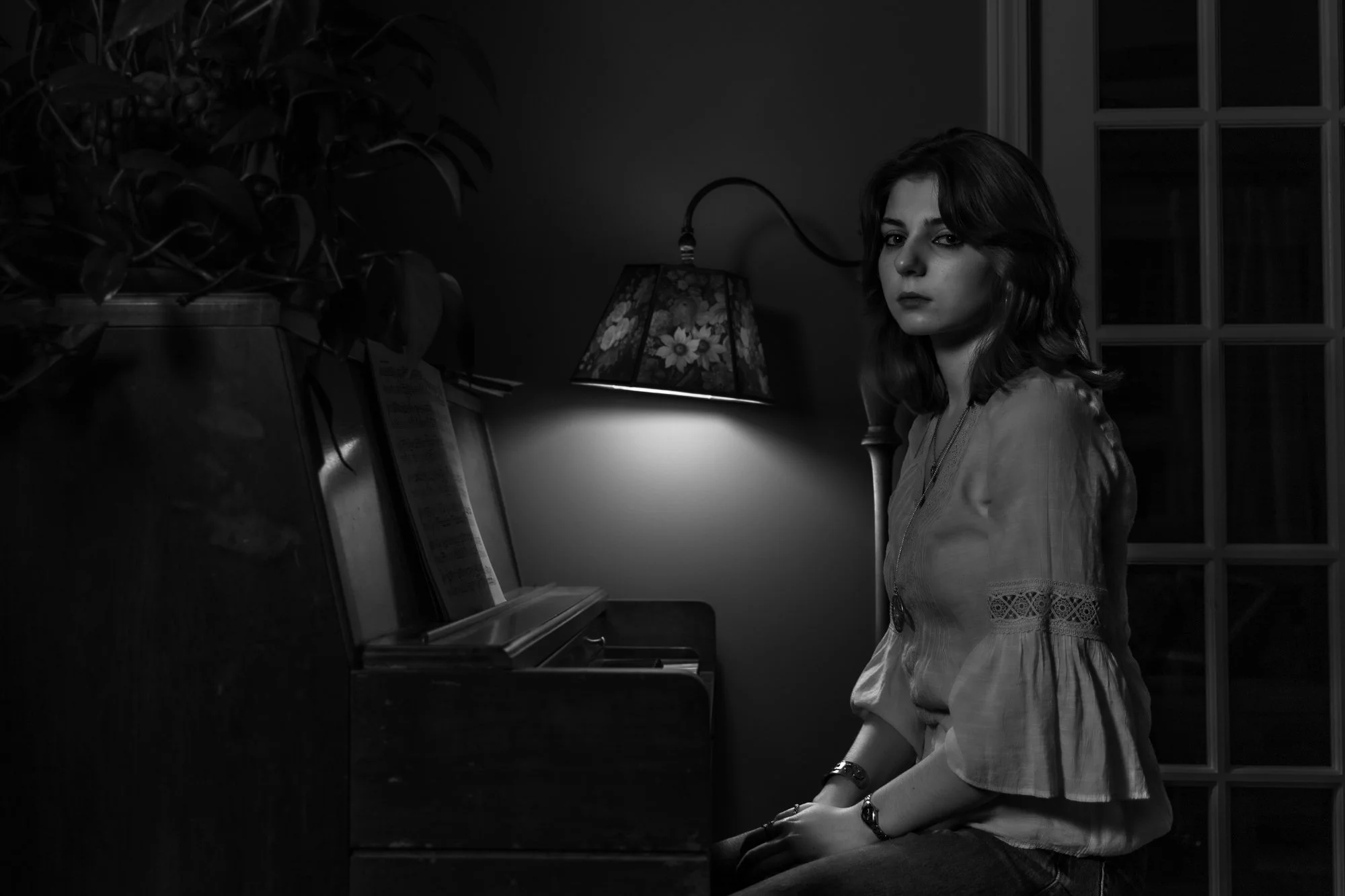 A woman with dark hair sitting at a piano in a dimly lit room, with a floral lampshade on a table behind her and a window in the background.