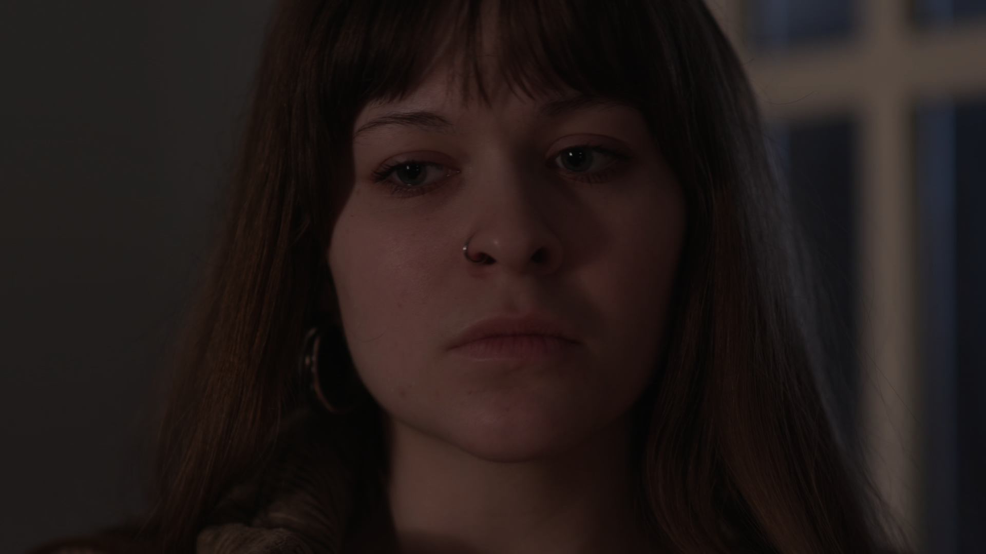 A young woman with long brown hair, a nose ring, and hoop earrings looking downward with a neutral expression.
