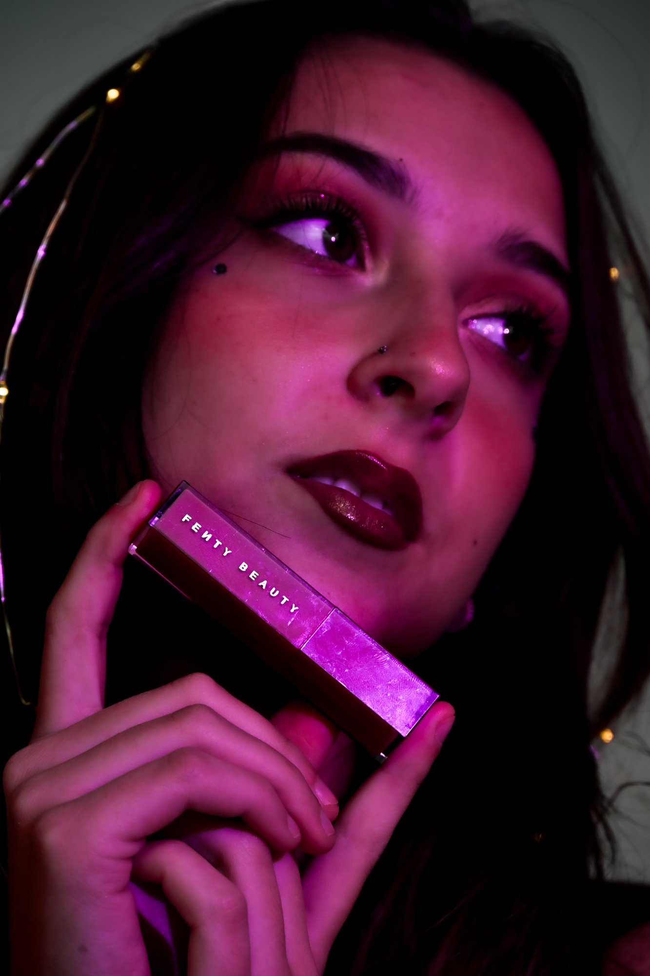 Close-up photograph of a woman with dark hair, wearing makeup, holding a pink box labeled 'FENTY BEAUTY,' with purple and pink lighting.