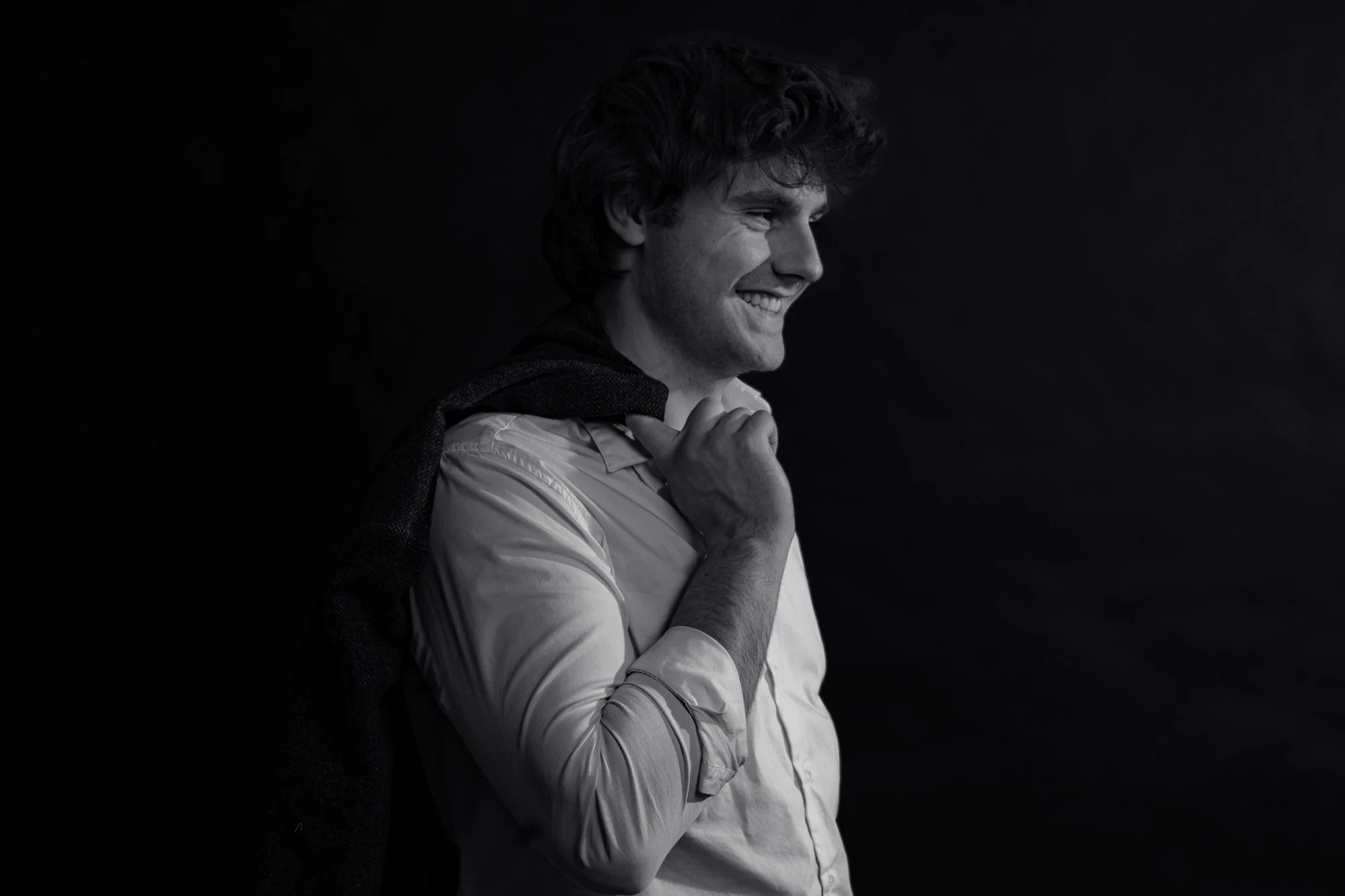 Black and white photo of a smiling man with curly hair, wearing a light shirt with a dark jacket draped over his shoulders, against a dark background.