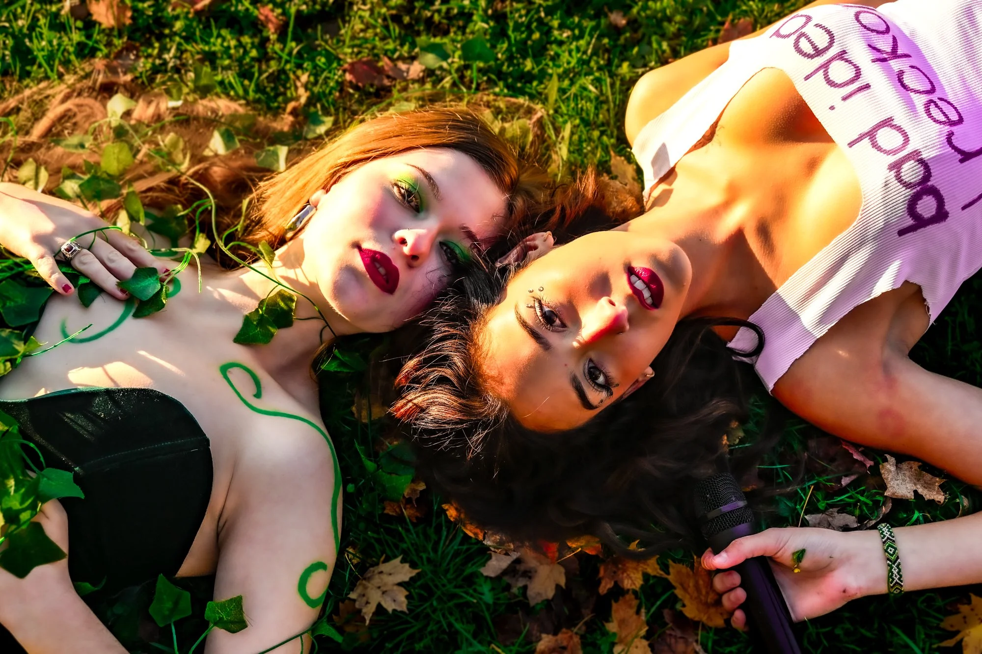 Two women lying on grass, surrounded by green leaves, with one holding a microphone. The women have vibrant makeup and are dressed in fashionable outfits, with one wearing a black top with cutouts and the other in a white shirt with writing.