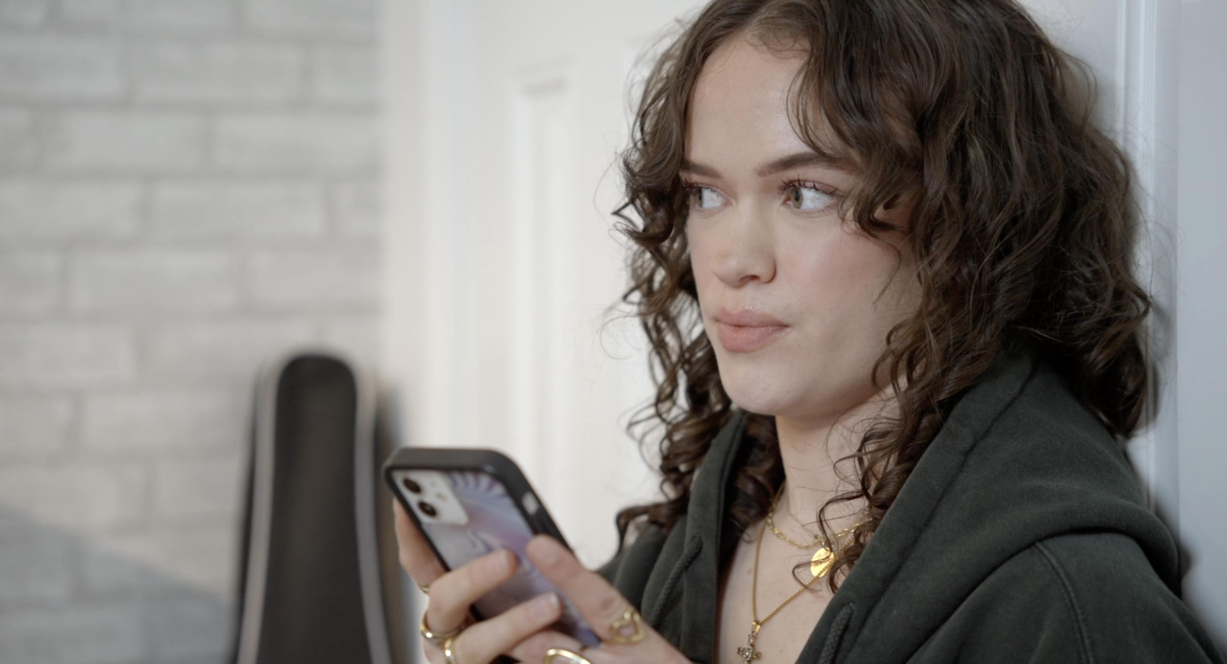 A young woman with curly brown hair, wearing a black jacket, holding a smartphone and looking to her left.