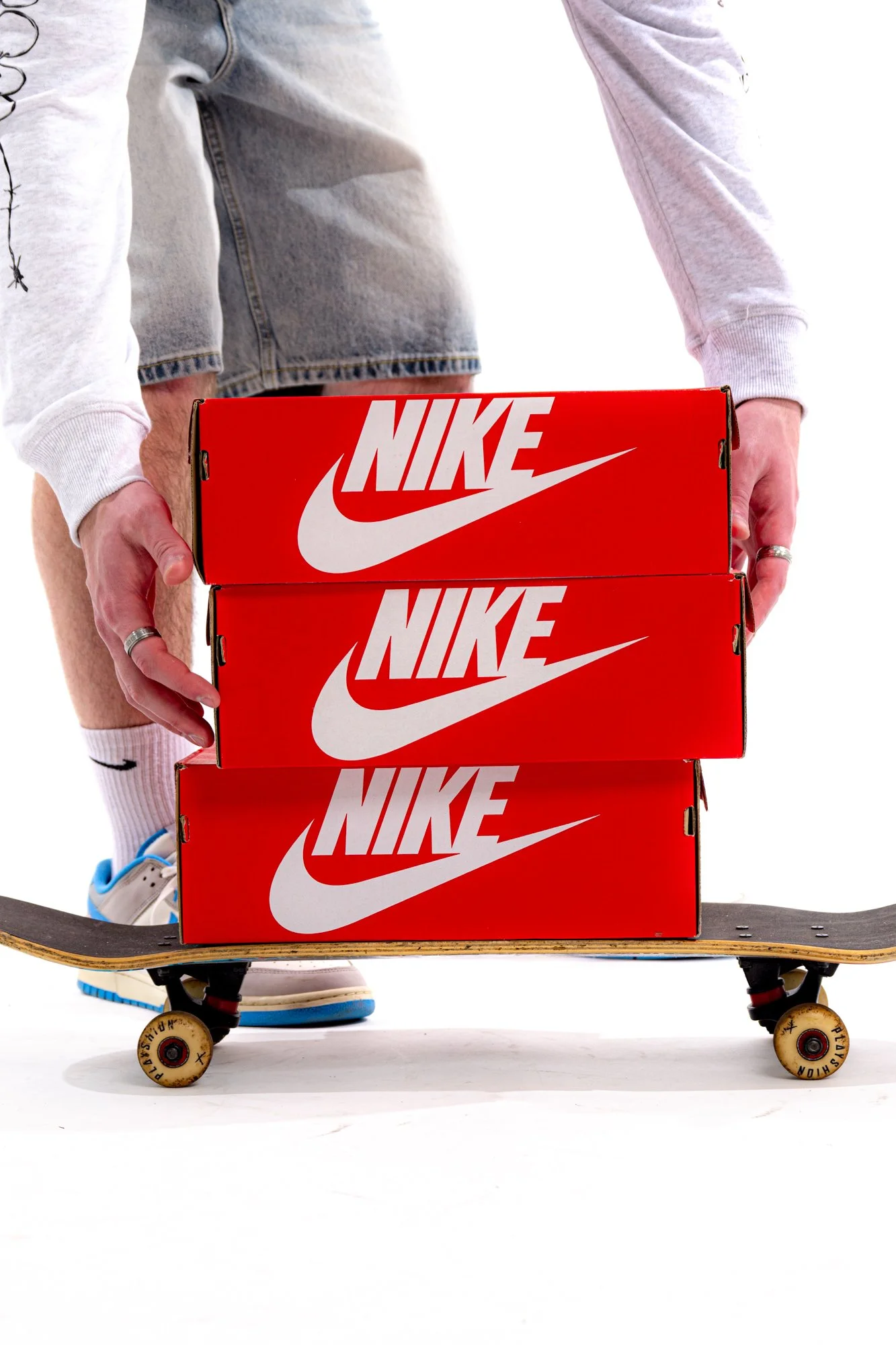 A person holding three red Nike shoeboxes on a skateboard, with skateboard wheels and part of their legs visible, wearing sneakers, white socks, and casual clothing.