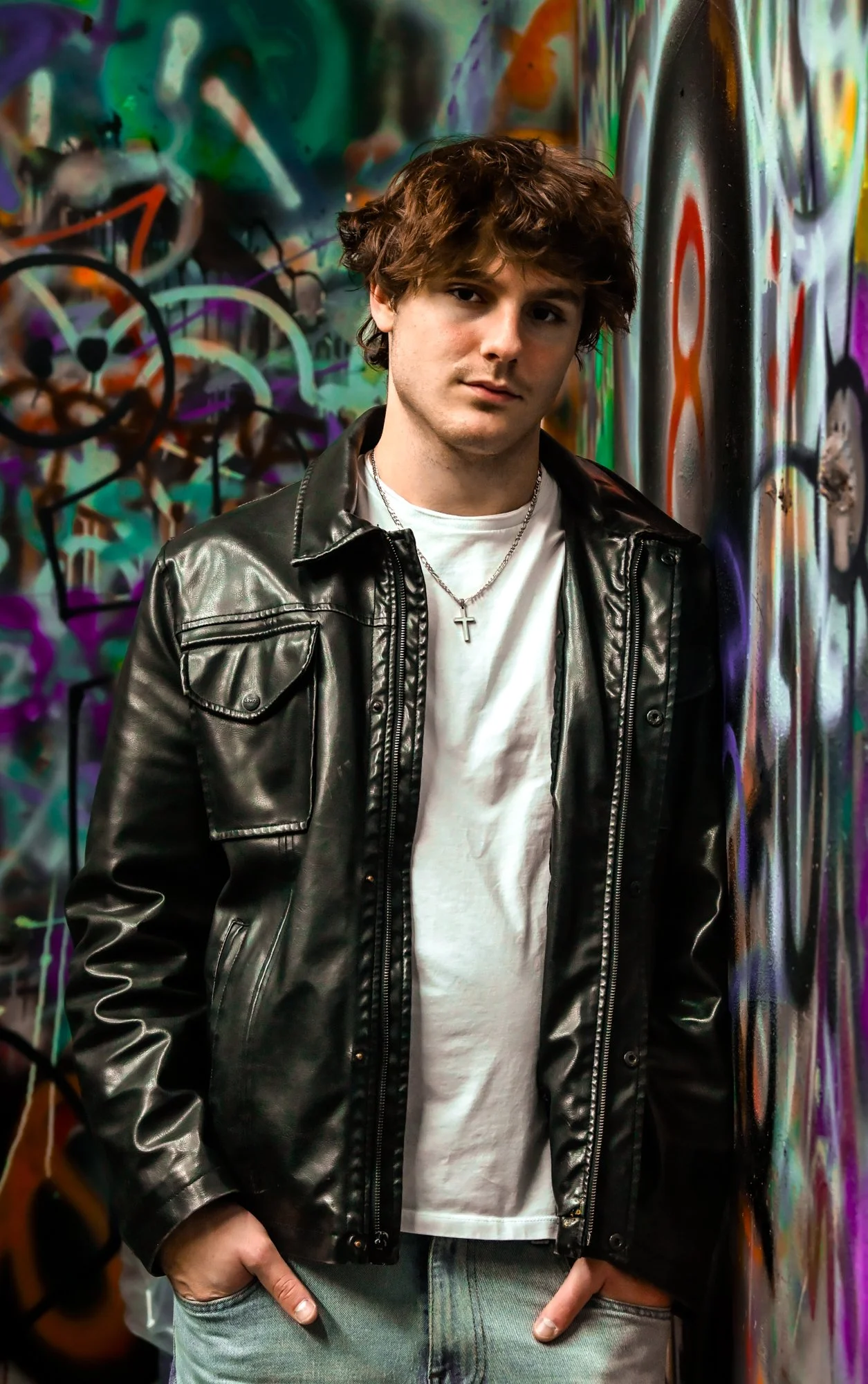 Young man in a black leather jacket and white t-shirt standing against a colorful graffiti wall.