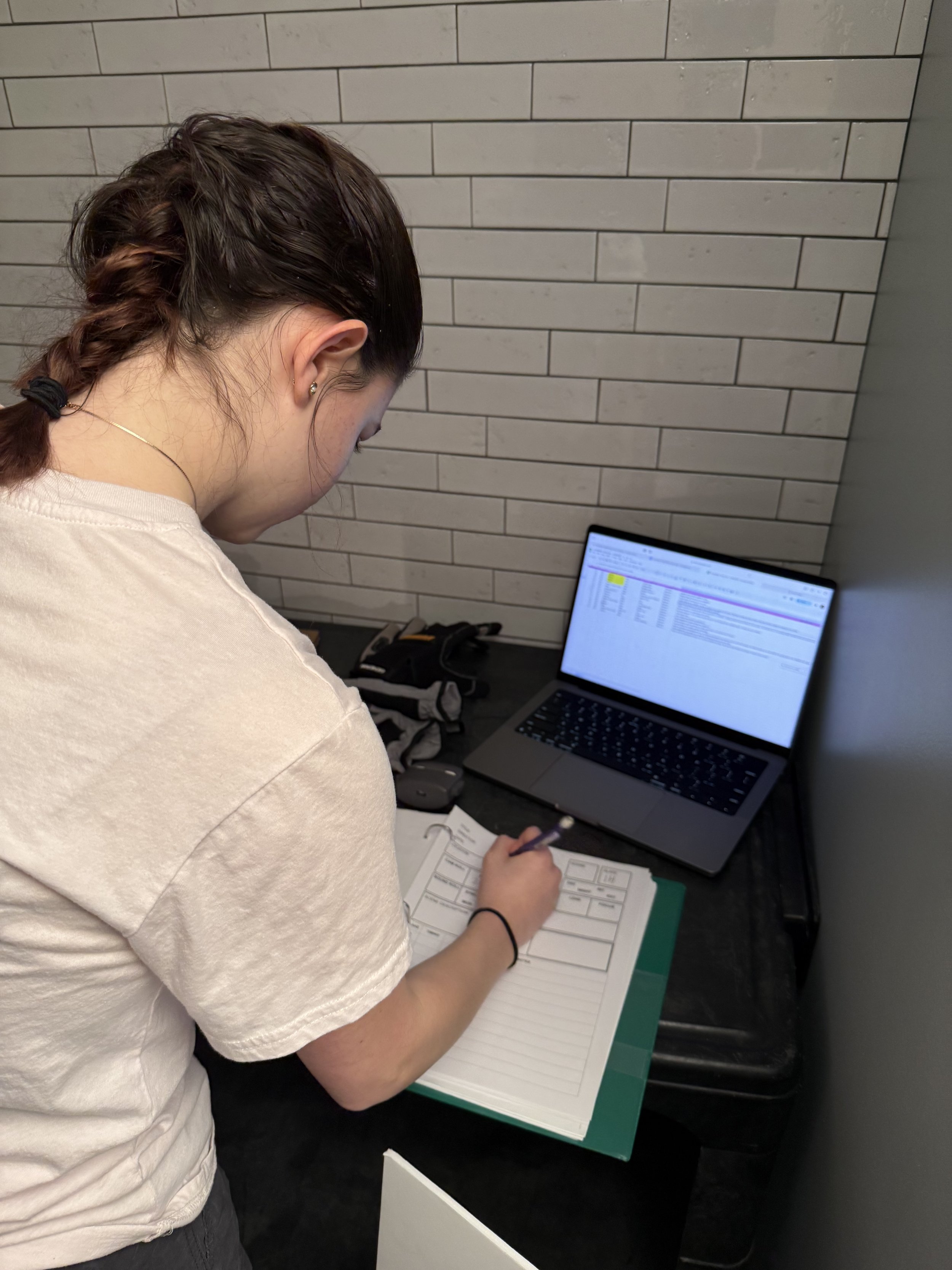 A woman with braided dark hair, wearing a white t-shirt, is sitting at a desk writing in a notebook. There is a laptop open in front of her displaying a spreadsheet, along with a mouse and some papers on the desk.