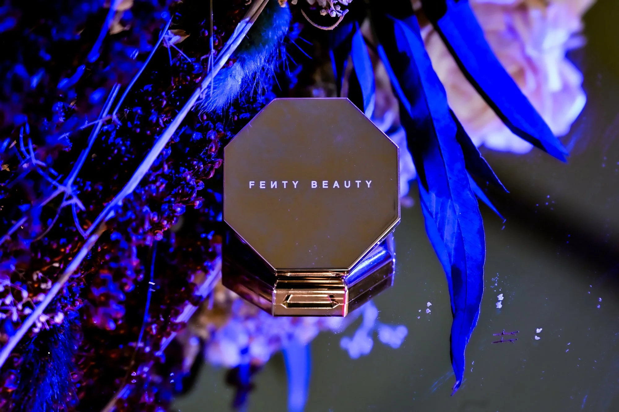 A reflective makeup compact with the words 'Fenty Beauty' on the cover, placed among purple flowers and blue-tinted leaves under vibrant lighting.