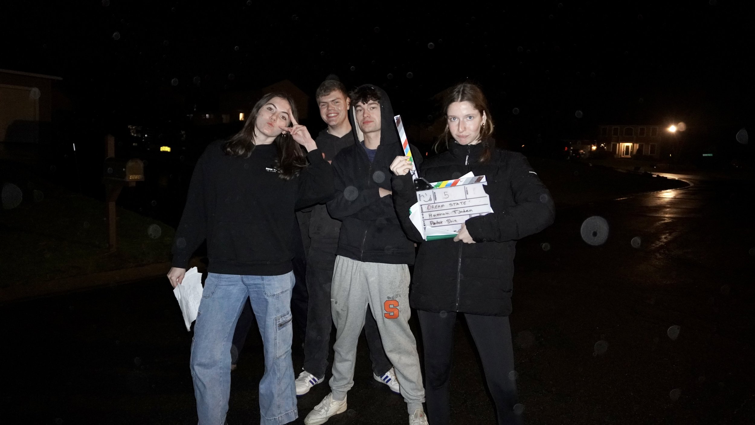 Four young people standing outdoors at night, two women and two men, one woman holding a clapperboard, and they appear to be filming or at a film set.