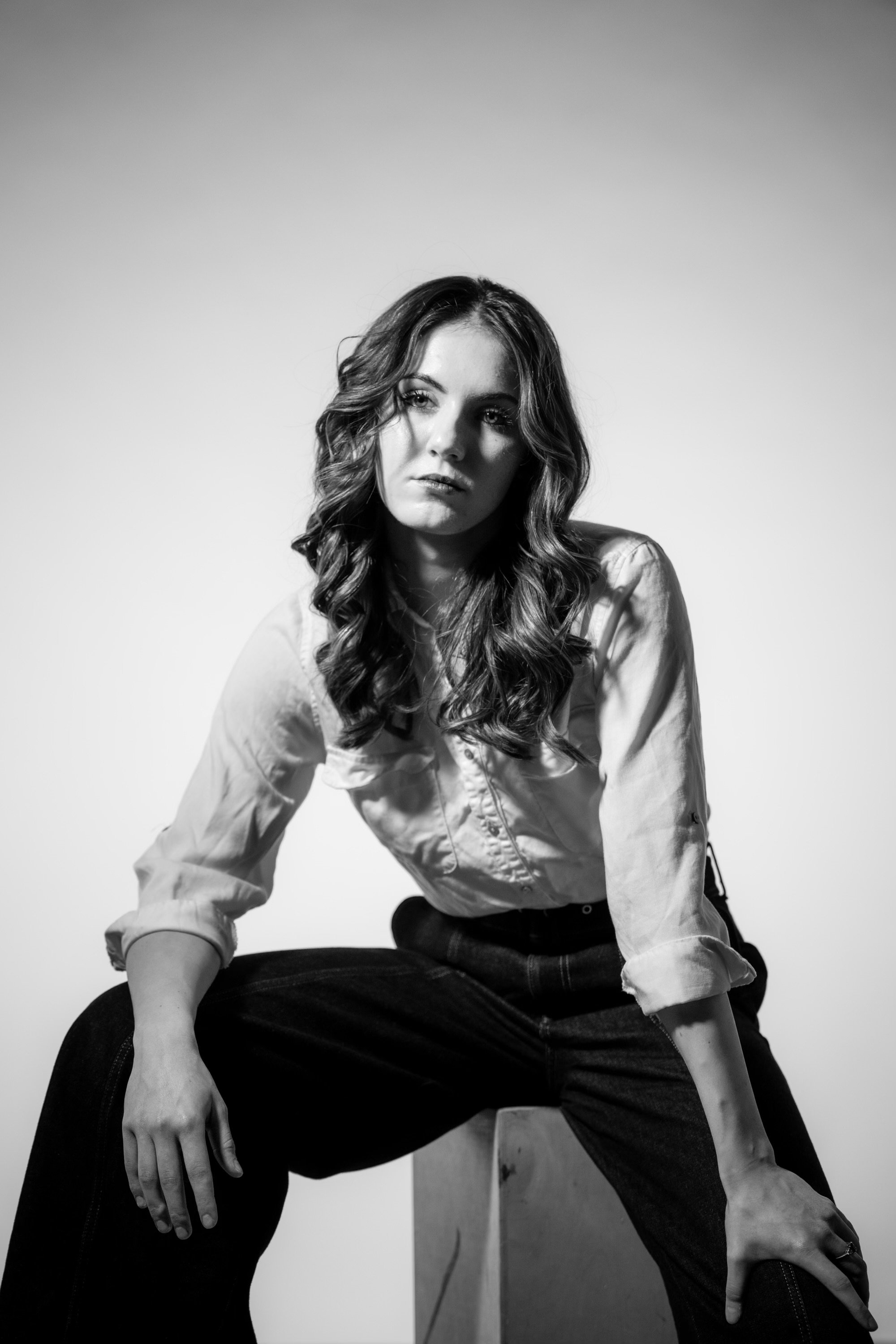 Black and white photo of a woman with long wavy hair, sitting on a stool, wearing a light-colored shirt and dark pants, against a plain light background.