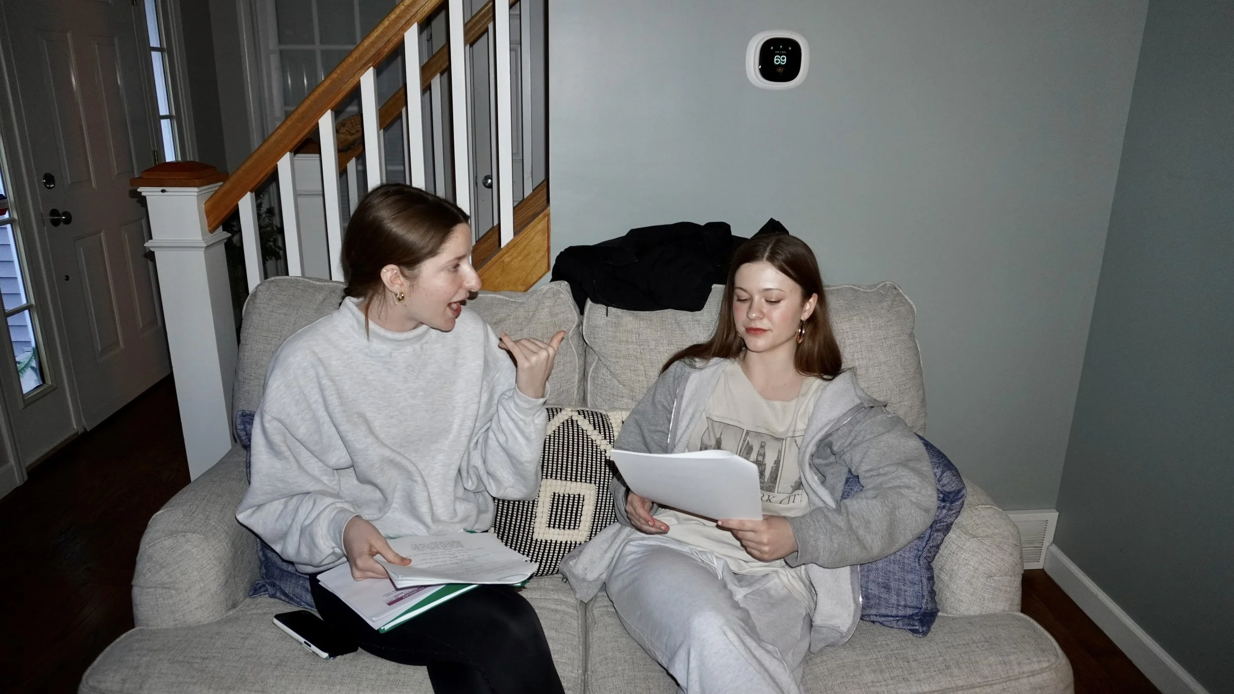Two young women sitting on a light-colored couch in a living room, looking at papers and a tablet, with one woman gesturing and talking while the other listens with a serious expression. A staircase and part of an entry door are visible in the backgr