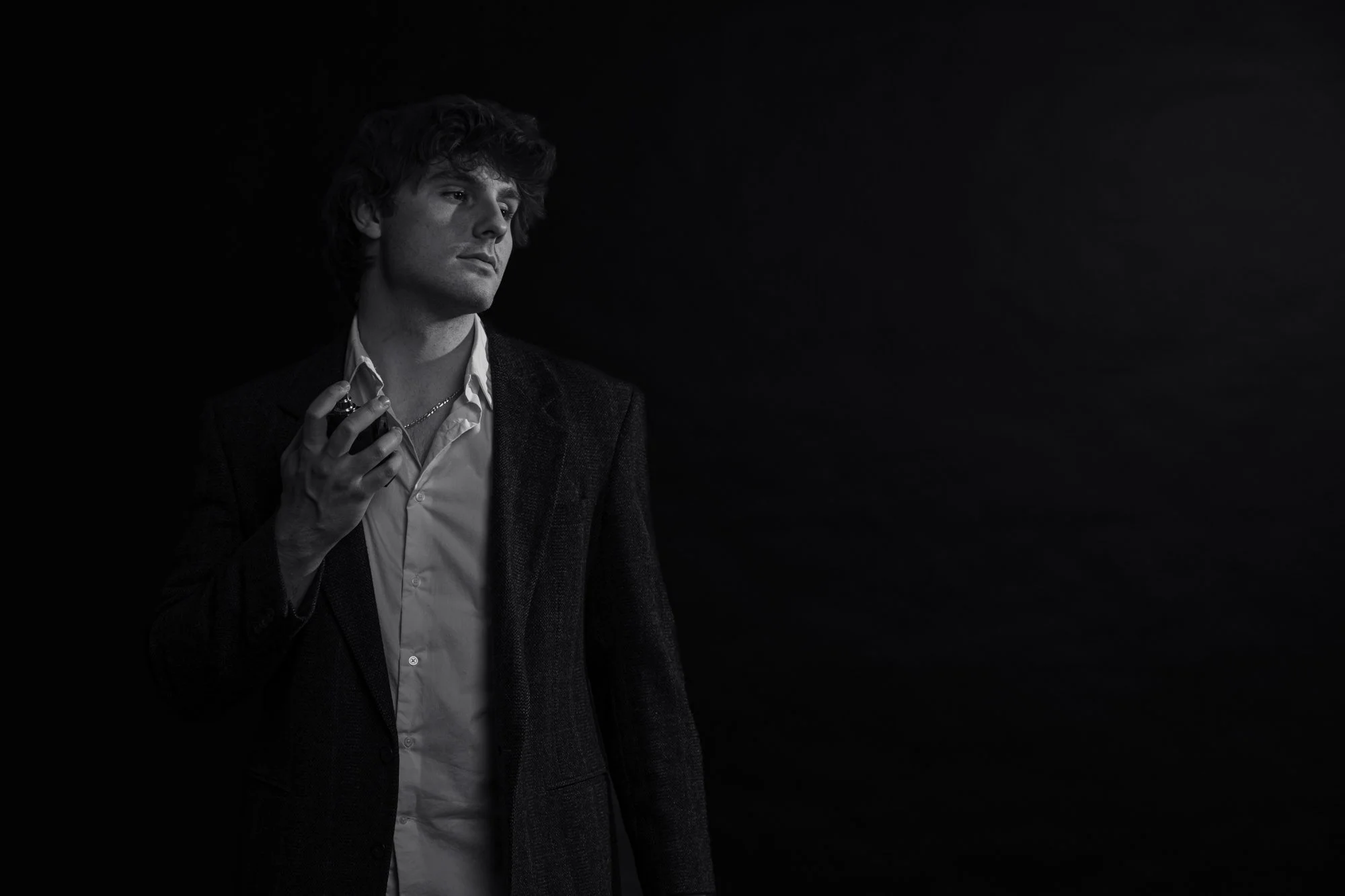 Black and white photo of a young man with curly hair wearing a blazer over a button-up shirt, holding sunglasses in his right hand, against a dark background.
