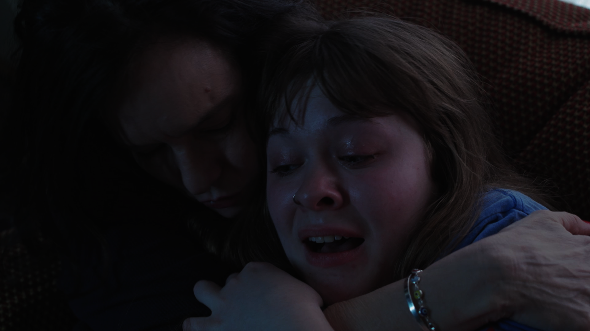 A woman and a young girl are embracing on a dark background. The girl appears to be crying, and the woman is holding her close.