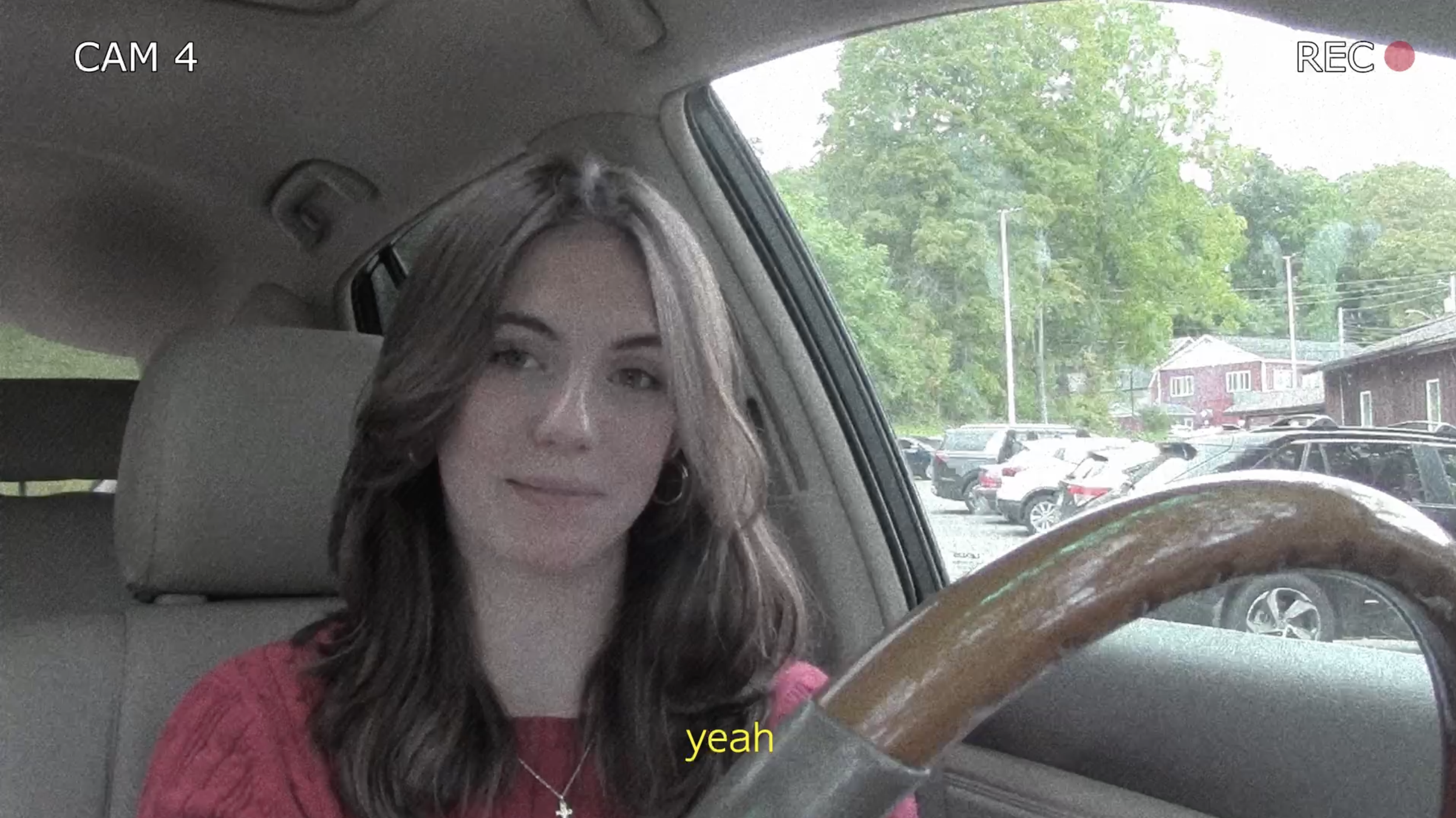 A woman with wavy brown hair and earrings sitting in the driver's seat of a car, wearing a pink sweater, with a parking lot, trees, and buildings visible outside the window.