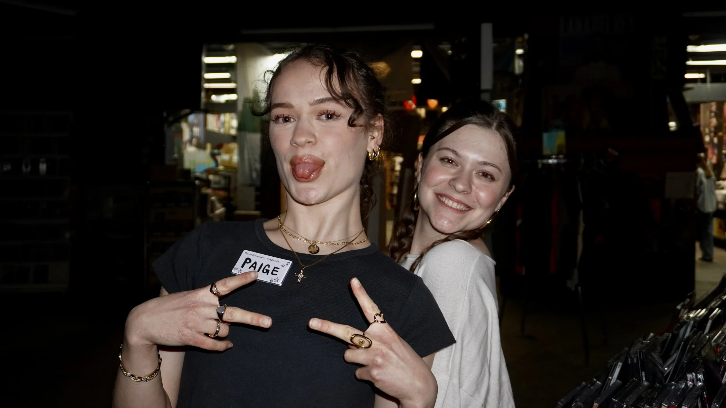 Two young women posing at an indoor event, one making a peace sign and sticking out her tongue, wearing a name tag that says "Paige".