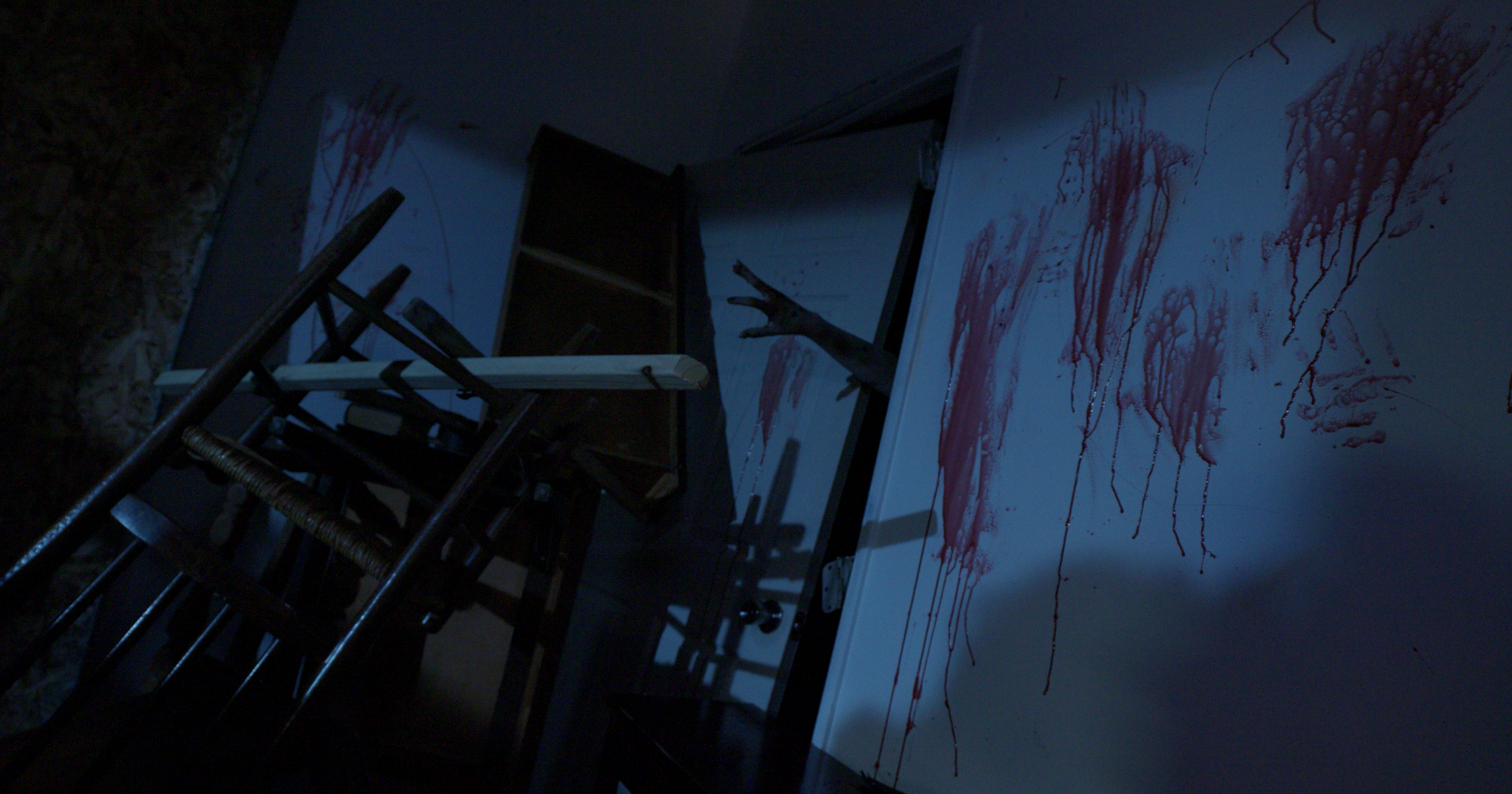 A room with a collapsed whiteboard covered in red paint or blood splatters, overturned chairs, and a person's arm reaching out from behind the whiteboard in a dimly lit environment.