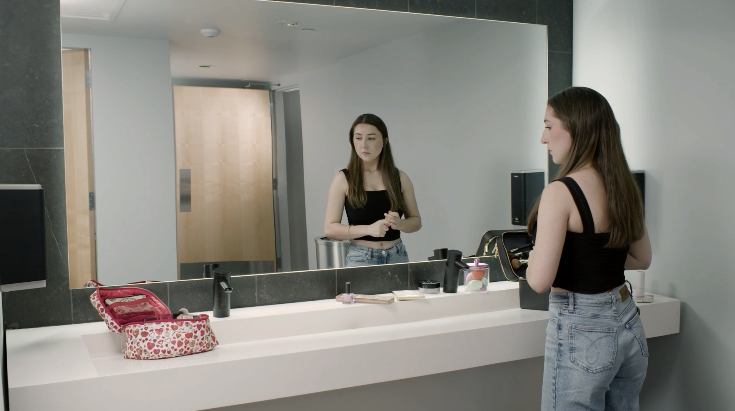 A young woman looking at herself in a mirror above a bathroom sink, with makeup items and a pink bag on the counter, in a modern bathroom.