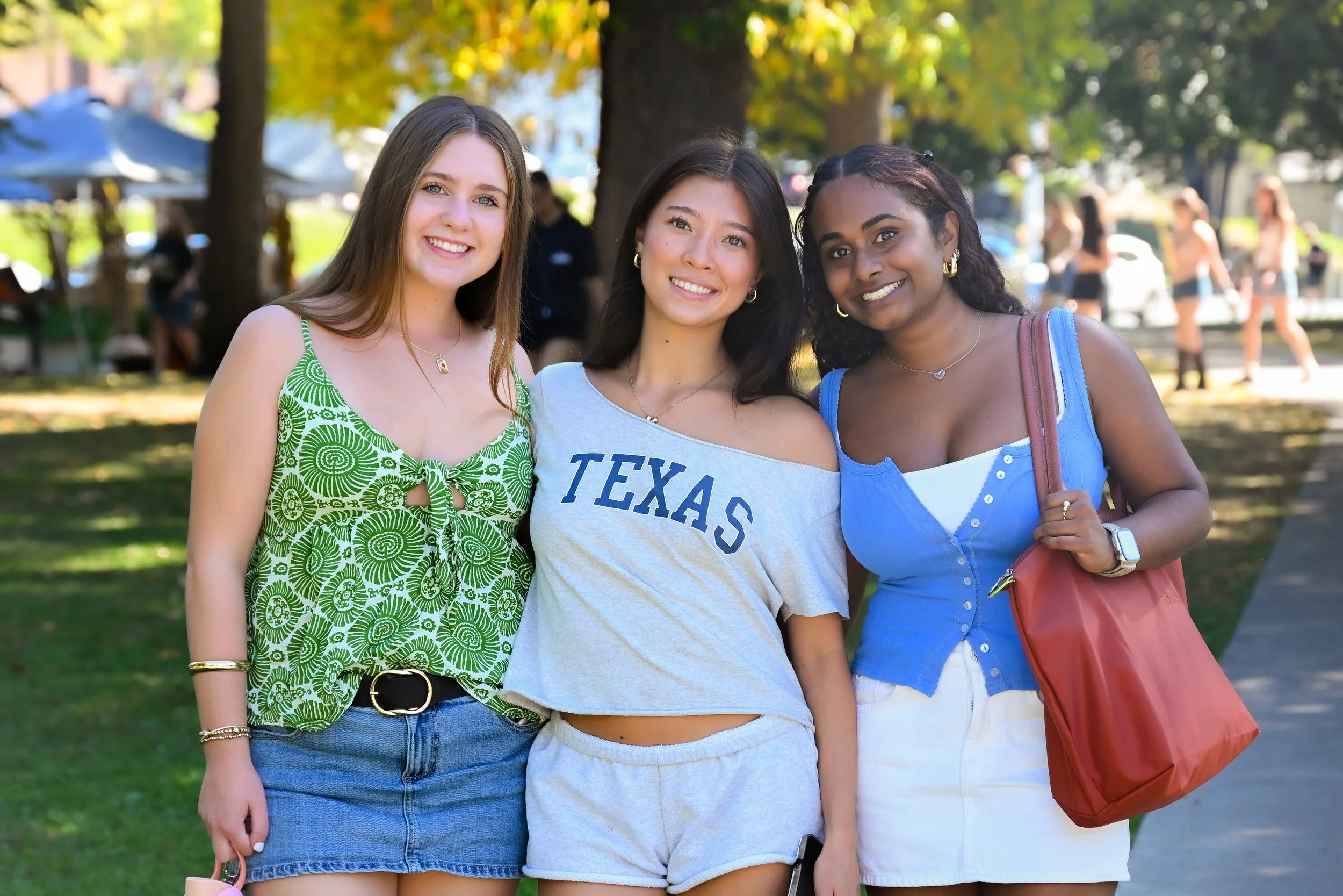 Three smiling young women standing together outdoors in a park with trees and people in the background. They are wearing casual summer clothes, with one in a green patterned top, one with a grey T-shirt that says 'TEXAS', and one in a blue sleeveless